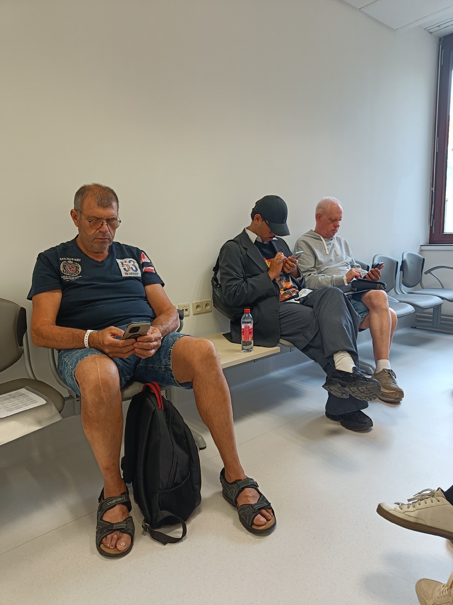 Three men are seated in a waiting room. The man on the left is wearing a dark blue t-shirt and denim shorts, and he is looking at his phone. He has glasses and short gray hair. The man in the middle is wearing a dark suit jacket over a t-shirt and dark pants, and he is also looking at his phone. He is wearing a black baseball cap. The man on the right is wearing a light gray hooded sweatshirt and denim shorts, and he is looking at his phone. He has short white hair. There are chairs behind the men, and a window is visible on the far right. The room appears to be a medical waiting area.