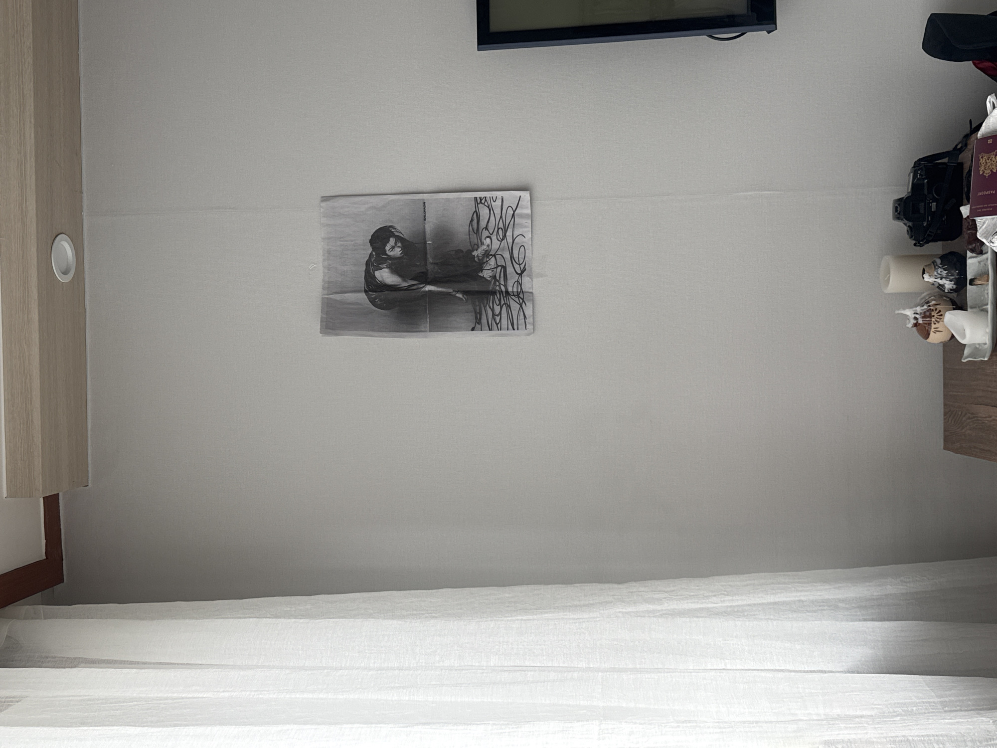 The image shows an interior space, possibly a bedroom, with a white wall and a white bed sheet. A black and white photograph is affixed to the wall. A flat-screen television is mounted near the top of the wall. A shelf to the right holds several items, including a camera, a passport, and decorative objects. A cabinet is partially visible on the left side of the frame. There is no indication of the location being in Seoul, South Korea.
