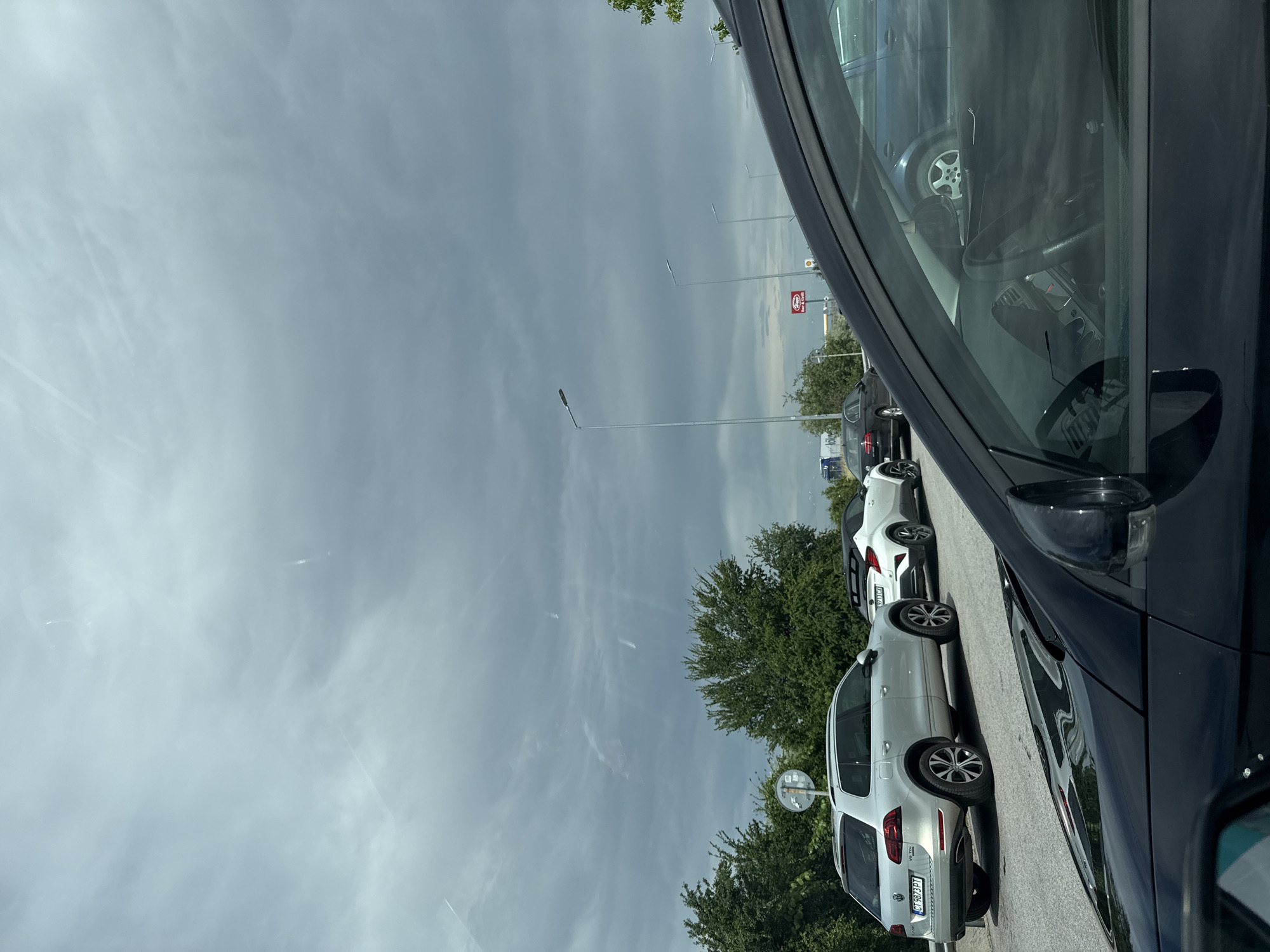 The image captures a daytime scene from within a dark-colored vehicle, likely parked, looking out over a parking lot or roadside area. The foreground on the right is dominated by the interior of the car, showing the steering wheel, dashboard, and the side mirror.
A line of cars is parked facing away from the viewer on the left side of the frame. The most prominent is a silver Volkswagen Tiguan SUV, clearly visible in the mid-ground. Its rear license plate, "CT 6873 PT," indicates a Bulgarian registration (Stara Zagora region), aligning with the provided location context of Radinovo, Bulgaria. Behind the silver SUV, a white SUV and another dark car are also parked.
The setting includes scattered green trees behind the parked vehicles and tall streetlights or lamp posts extending into the distance. A circular convex mirror on a pole is visible near the silver SUV. In the far background, a red and white sign, possibly for a gas station or shop, can be seen, though the text is indistinct due to distance. A white truck with a blue logo is partially visible beyond the sign.
The sky is partly cloudy and bright, suggesting a clear day with some atmospheric haze or wispy clouds. The overall impression is that of a stop in a commercial or transit area. No people are visible, and no specific event or activity other than parking is taking place.
