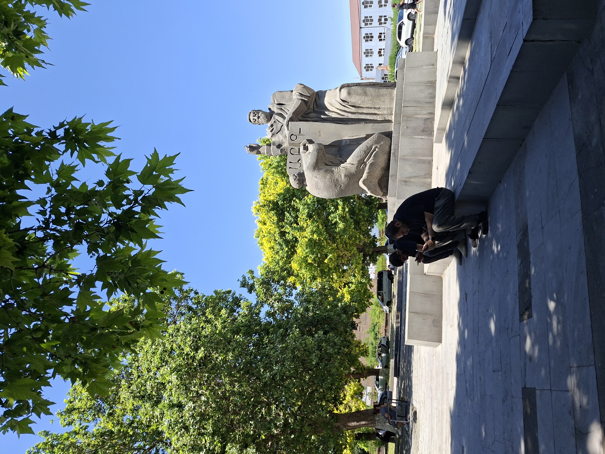 In this outdoor scene, a stone monument depicting two figures stands prominently under a bright blue sky. The larger figure, seated, is an elderly man with a beard, wearing draped robes and holding a book or tablet. To his left, a kneeling figure appears to be receiving something or looking up at the seated figure. The monument is situated on a tiered stone platform in what seems to be a public space, possibly a park or square.
In the foreground, three men are seated on a concrete ledge, each engrossed in their mobile phones. They are casually dressed, with two in dark shirts and one in a lighter shirt. The ground around the monument and the seating area is paved with stone tiles.
Lush green trees with full foliage frame the top and left side of the image, with some dappled sunlight filtering through their leaves. In the background, a modern building with a light-colored facade and numerous square windows is visible, along with several parked cars. The overall atmosphere suggests a warm, sunny day in a city setting.