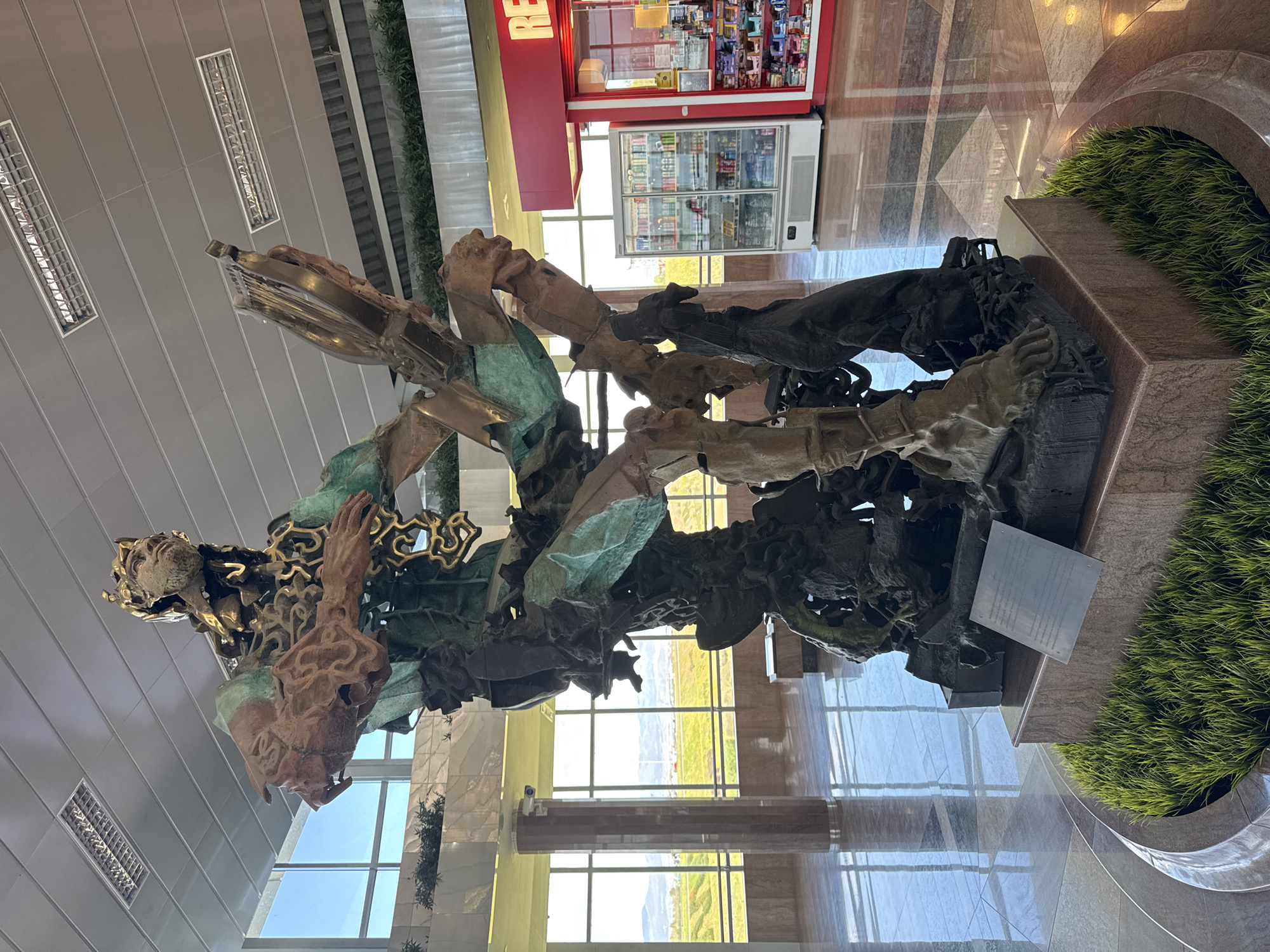 The image captures a large, intricate metal sculpture located in what appears to be a modern building, likely an airport or a large public space. The sculpture features figures and abstract shapes, with some elements showing a greenish patina, suggesting age and exposure to the elements. The base of the sculpture is surrounded by decorative landscaping. A vending machine can be seen in the background, further supporting the impression of a public area. Considering the location is Sofia, Bulgaria, it is highly probable that the sculpture is located at Sofia Airport. The polished floor and large windows suggest a modern, well-maintained environment.