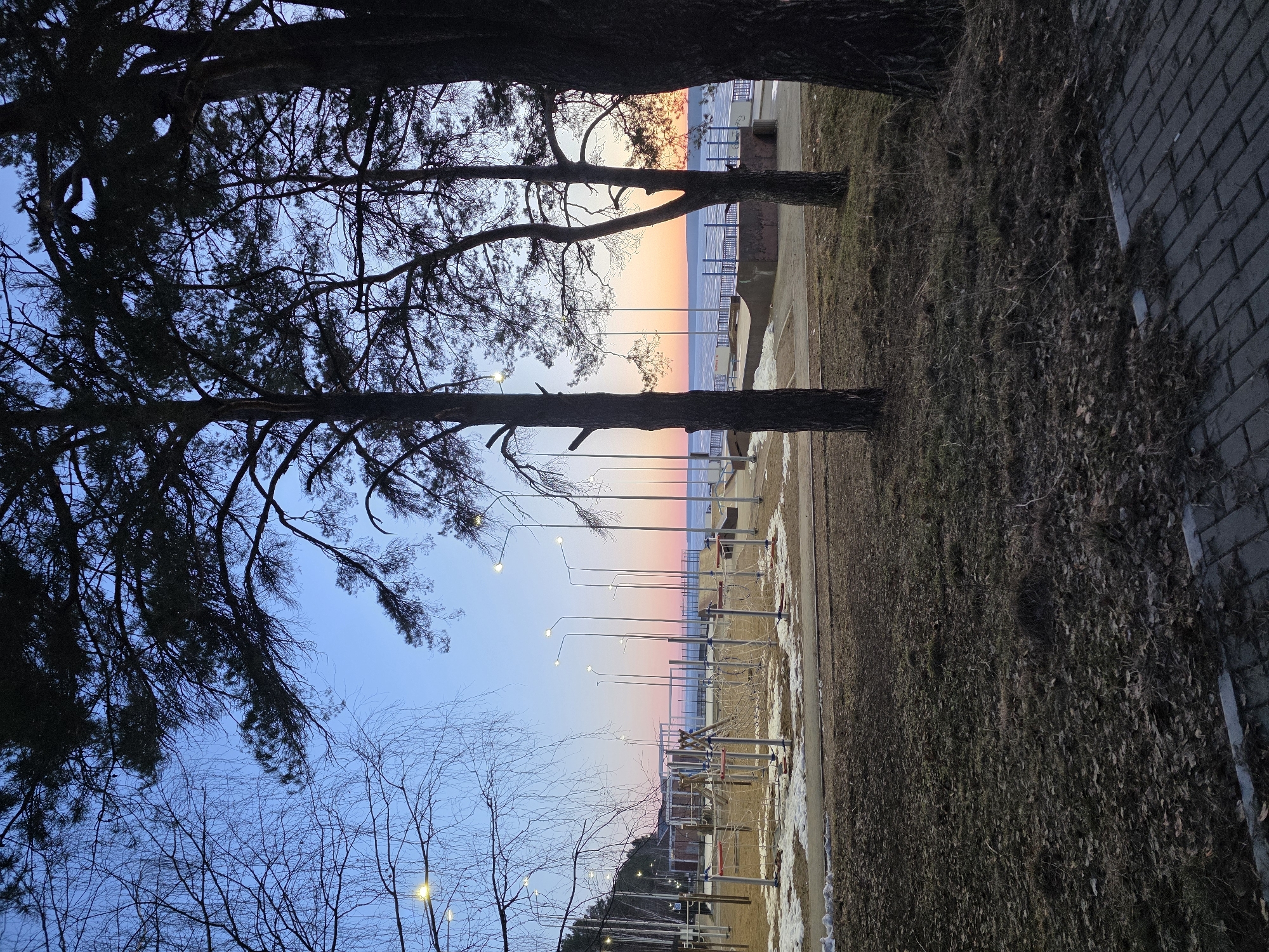 The image shows a view towards the Kama River from the pedestrian zone in the city of Chaikovsky, Perm Krai, Russia. A path runs along the riverbank, lined with exercise equipment and lighting poles. Bare trees and pine trees are visible along the shoreline. The sky shows a gradient of colors from blue to orange, indicating either sunrise or sunset. The ground has patches of snow and dry vegetation. A brick pavement is seen to the right of the path. The scene appears to be deserted at the time of the photograph.