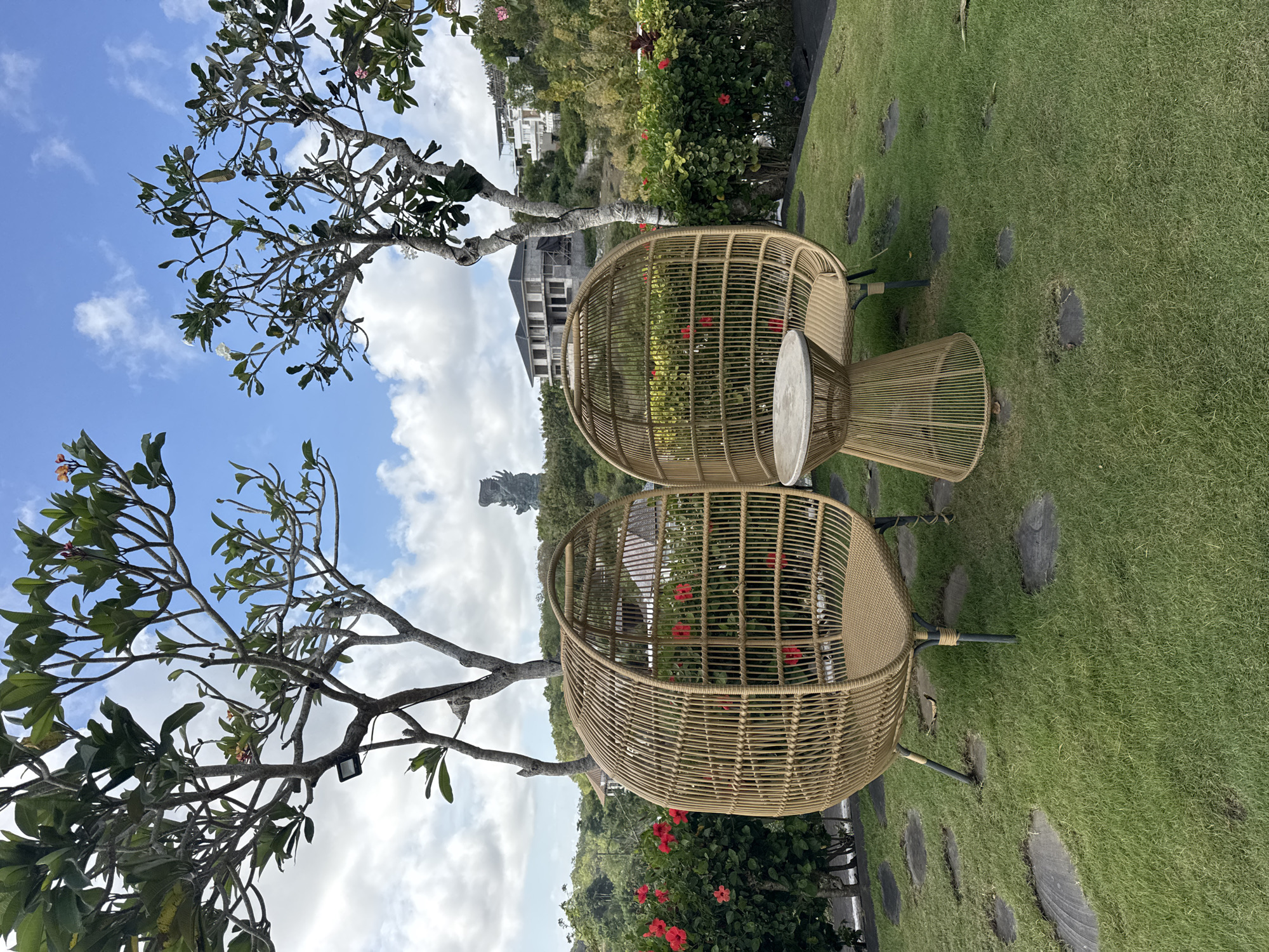 The image depicts a pair of wicker egg chairs and a small table set on a green lawn dotted with stone pavers. Overhead, a tree with sparse foliage extends into a blue sky with scattered clouds. In the background, buildings and greenery are visible, suggesting a residential or resort area. The statue of Garuda Wisnu Kencana is visible in the distance, indicating the location is in Kuta Selatan, Indonesia. The overall scene is peaceful and evokes a sense of relaxation.