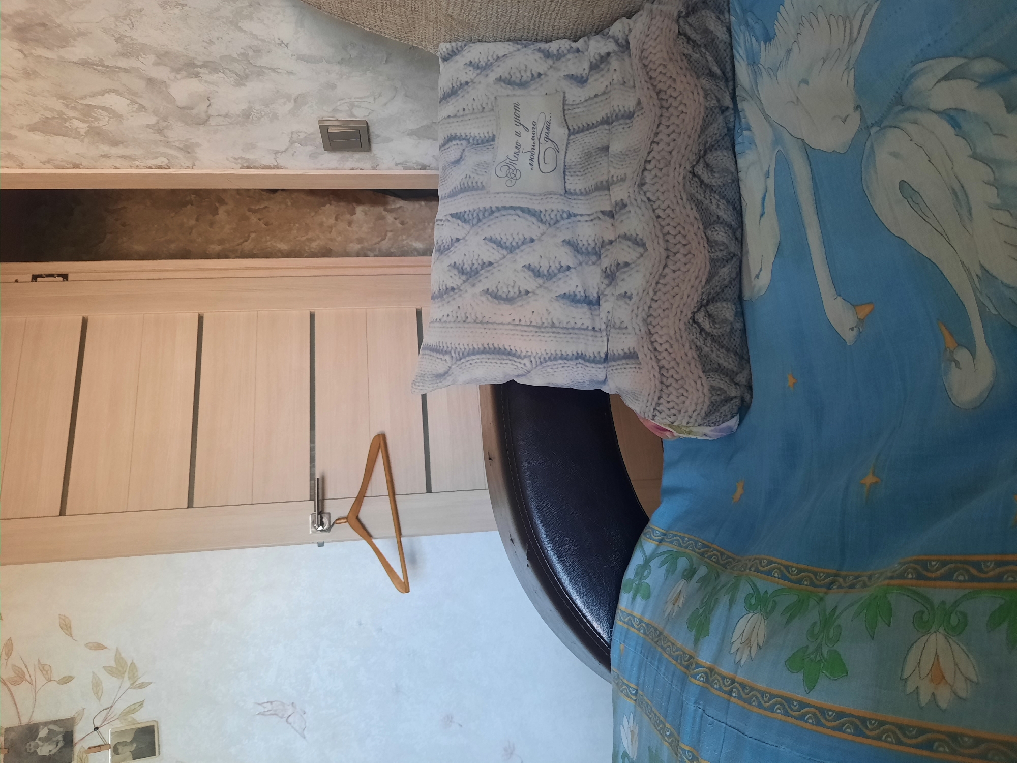 The image depicts an indoor scene, likely a living room or bedroom, with a focus on a blue bedspread featuring a swan pattern. A white knit pillow with a decorative inscription rests on the bedspread. In the background, a light-colored wooden door with horizontal paneling is visible. A wooden hanger hangs on a hook attached to the door. To the left of the door, a wall with a floral wallpaper design and two framed photographs is present. A light switch is mounted on the wall to the right of the door. The scene is lit by an unseen light source, casting a soft glow on the objects.