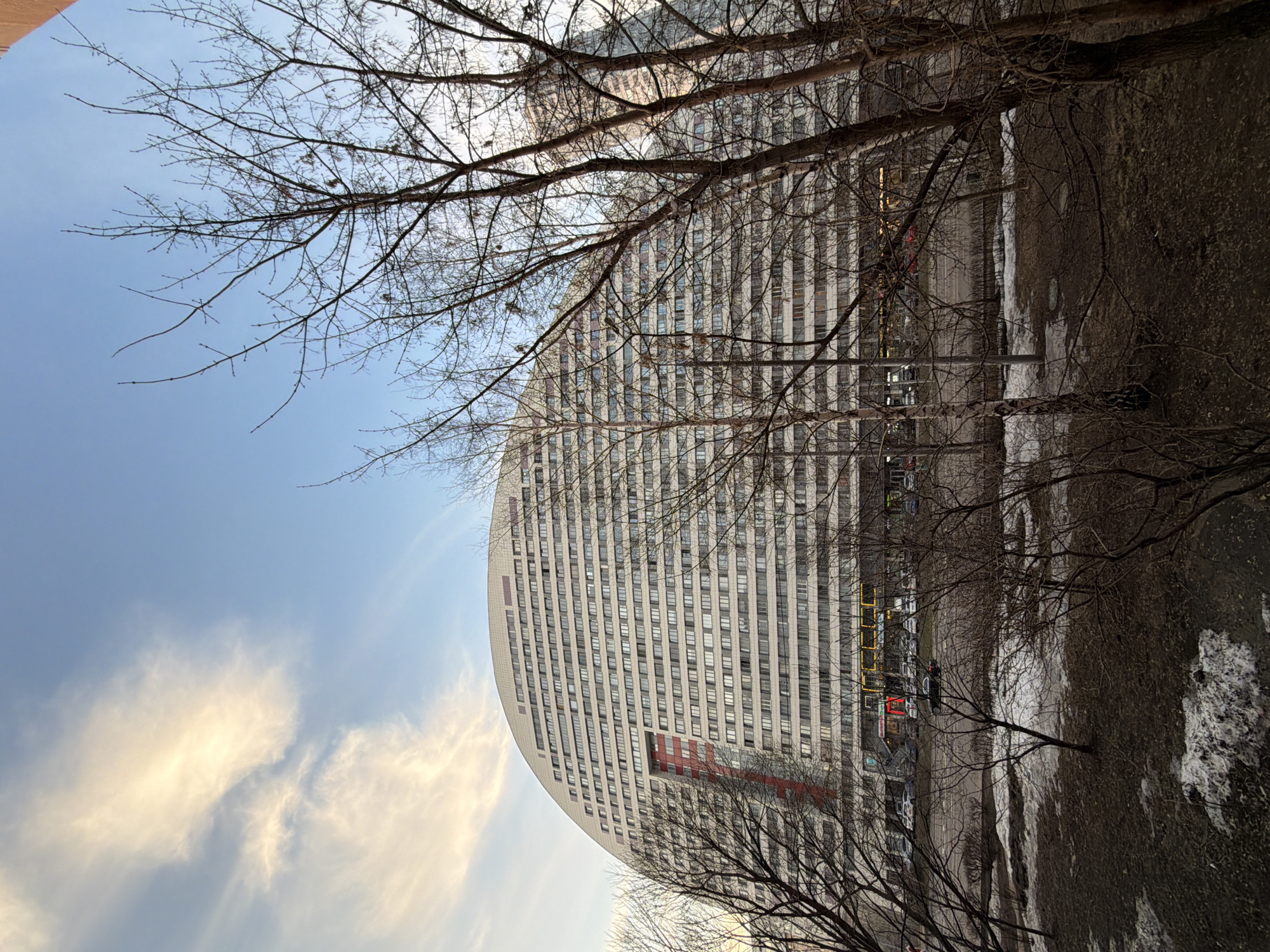 The image depicts an urban landscape in Khimki, Russia, featuring the distinctive multi-story, semi-circular building at Ulitsa Zelenskogo, 16. The scene is captured during daylight, likely late winter or early spring, as evidenced by patches of melting snow and bare deciduous trees lacking foliage.
The dominant subject is a massive, curved building, predominantly light grey or white, with a convex facade composed of numerous uniform rectangular windows arranged in horizontal rows. A prominent, irregular vertical red architectural feature is integrated into the left side of the building's visible curve. The ground level of the building reveals commercial establishments with illuminated signage, featuring yellow and red lighting.
In the mid-ground, a road or parking area runs horizontally, with multiple vehicles parked along its edge. One dark-colored sedan is observed in motion on this road. The foreground consists of unpaved, uneven ground covered with dark earth, sparse dry vegetation, and scattered patches of white snow. Numerous bare deciduous trees, characterized by thin branches, are distributed across the foreground and mid-ground, partially obstructing the view of the building. The sky above is a light blue with scattered, wispy white clouds, illuminated by natural sunlight. No individuals are clearly discernible, and no specific actions or interactions are visible.
