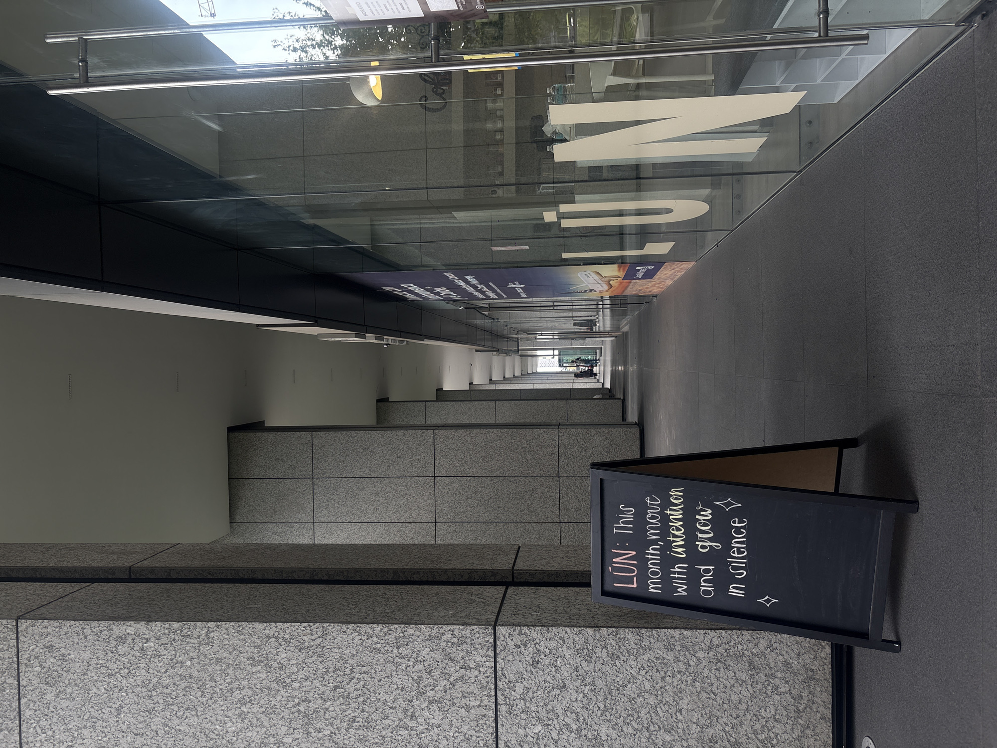 A photograph shows a long, narrow passage in Mexico City, Mexico. The camera's perspective is oriented along the corridor's axis, enhancing the sense of depth.
On the right side, a black sandwich board stands. The text on the board reads "LŪN: This month, move with intention and grow in silence."
The corridor is framed by glass walls on the upper left and right sides. Reflected within the glass are partial words "Cafe", "HORARIO", and the large word "MUSEO". The reflections imply the corridor is adjacent to or connects to a museum.
The walls are constructed of square grey stone blocks. The ground is also stone, matching the walls.
In the far distance, light enters the corridor, illuminating the end of the passage. Silhouettes of people are faintly visible at the end of the hallway. The passage appears to be outdoor or semi-outdoor due to the ample natural light.