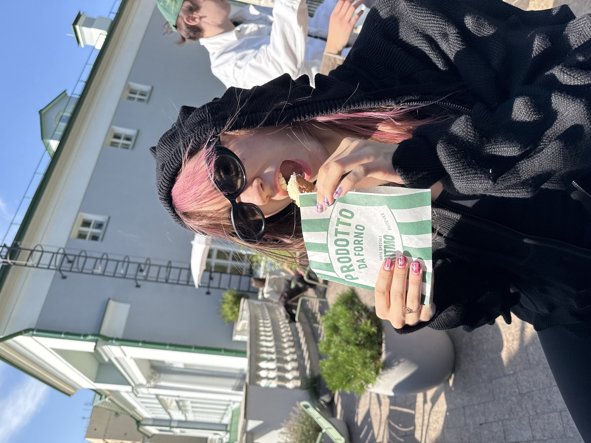A young woman with pink-dyed hair and sunglasses is eating a pastry from a green and white striped paper bag in an outdoor cafe setting. She is wearing a black hooded jacket. In the background, another person wearing a white jacket and green cap is visible. The cafe appears to be located in Moscow, Russia, indicated by the distinctive architectural style of the building behind the woman, which features a light blue facade, white trim, and classical columns, characteristic of many historical buildings in the city. The presence of a ladder on the side of the building is also a notable detail of the urban environment. The ground is paved with grey stones, and there are potted plants, suggesting a pleasant outdoor seating area. The weather is sunny with a clear blue sky.