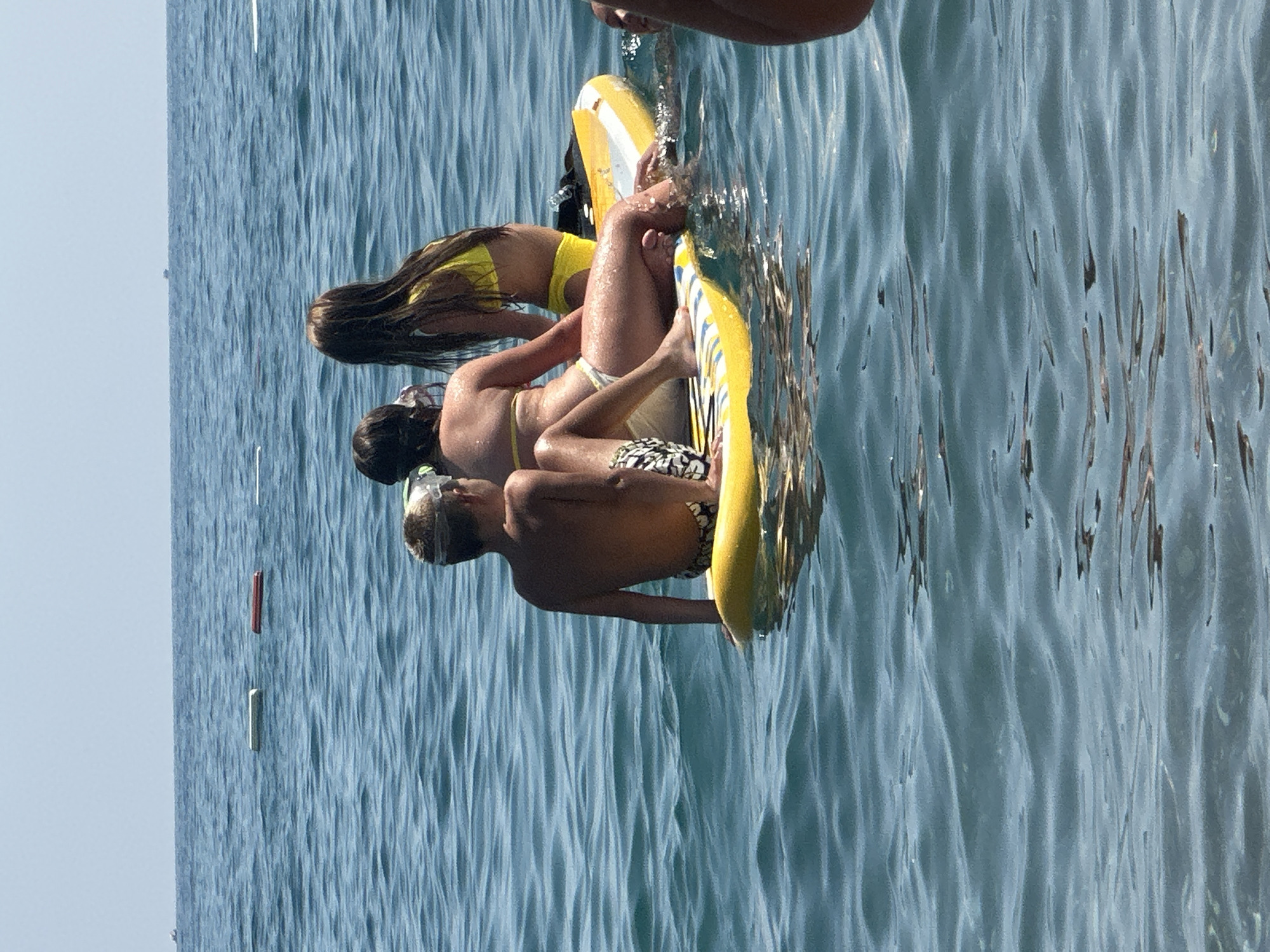 The image shows three people in the water, appearing to be a family enjoying a day at the beach. The water is a clear blue, with gentle waves suggesting a calm day. One person, possibly an adult, is partially visible at the top of the frame, with their hands reaching towards the water. Below them, two children are seated on a yellow and white inflatable mat. The child on the left, with long dark hair, is wearing a yellow bikini top. The child on the right, a boy, is wearing swimming trunks and has a snorkel mask around his neck. They are both positioned on the inflatable mat, which is partially submerged in the water.
The scene suggests a leisurely activity on a sunny day, likely during the summer. The bright sunlight reflects off the water, creating a shimmering effect. The presence of the inflatable mat indicates a recreational outing, possibly a swim or a playful session in the sea. The clear water and the relaxed postures of the individuals convey a sense of enjoyment and relaxation. In the background, a few vertical markers, likely buoys, are visible in the water, suggesting a designated swimming area. The horizon line is visible, with a clear, pale blue sky. The overall mood is peaceful and summery. No visible text is present in the image.