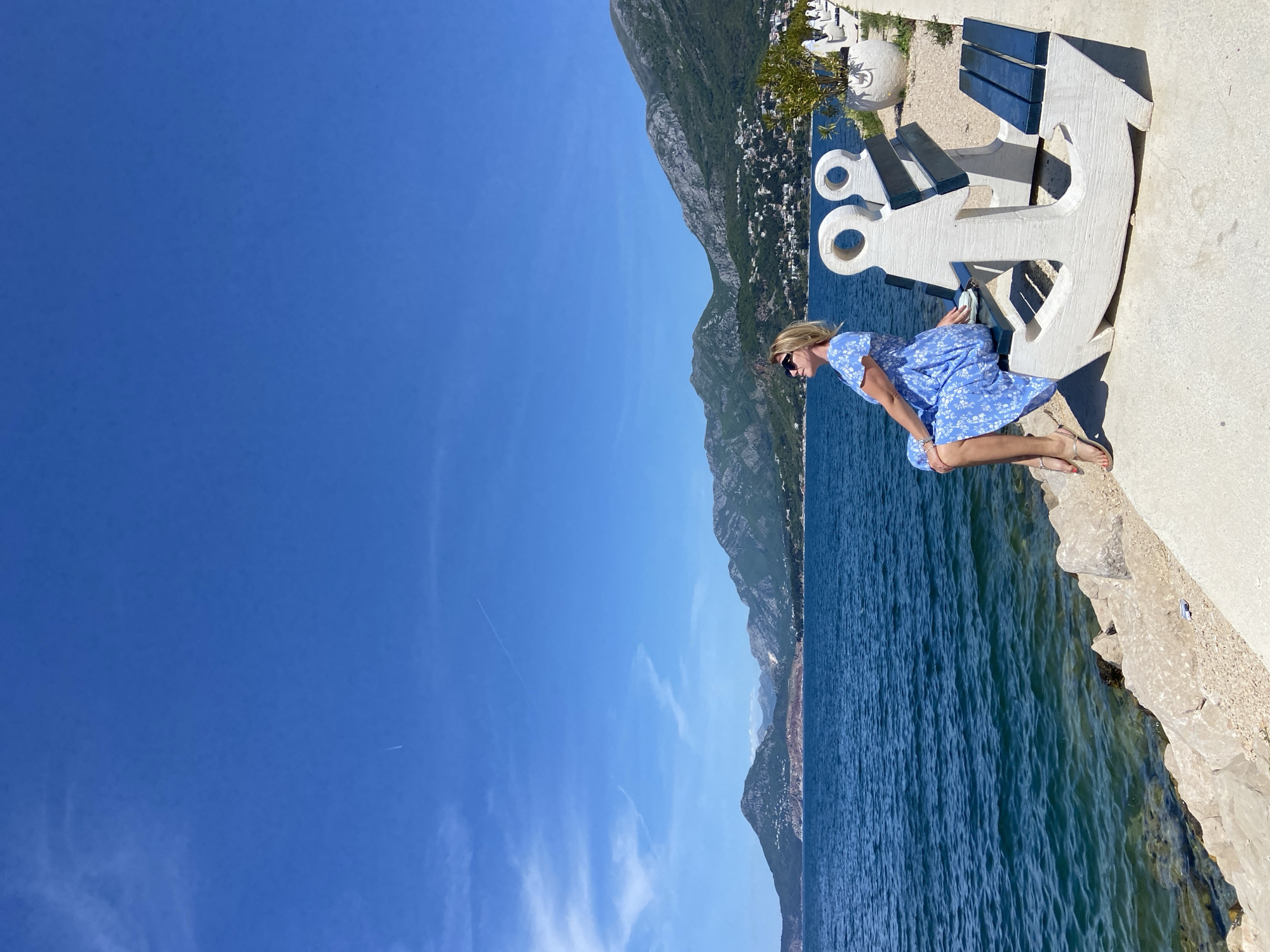 The image depicts a sunny outdoor scene featuring a woman seated on an anchor-shaped bench, seemingly a part of a larger structure. The background includes a body of water, presumably the sea, with mountains visible in the distance.
The woman is wearing a blue floral dress, sunglasses, and sandals. She is seated on the edge of the structure, her legs dangling over the side.
The anchor-shaped bench is made of a light-colored material and appears to be part of a larger concrete structure.
The location is most likely near the coast of the city of Bar, Montenegro, given the presence of the sea and mountainous terrain.