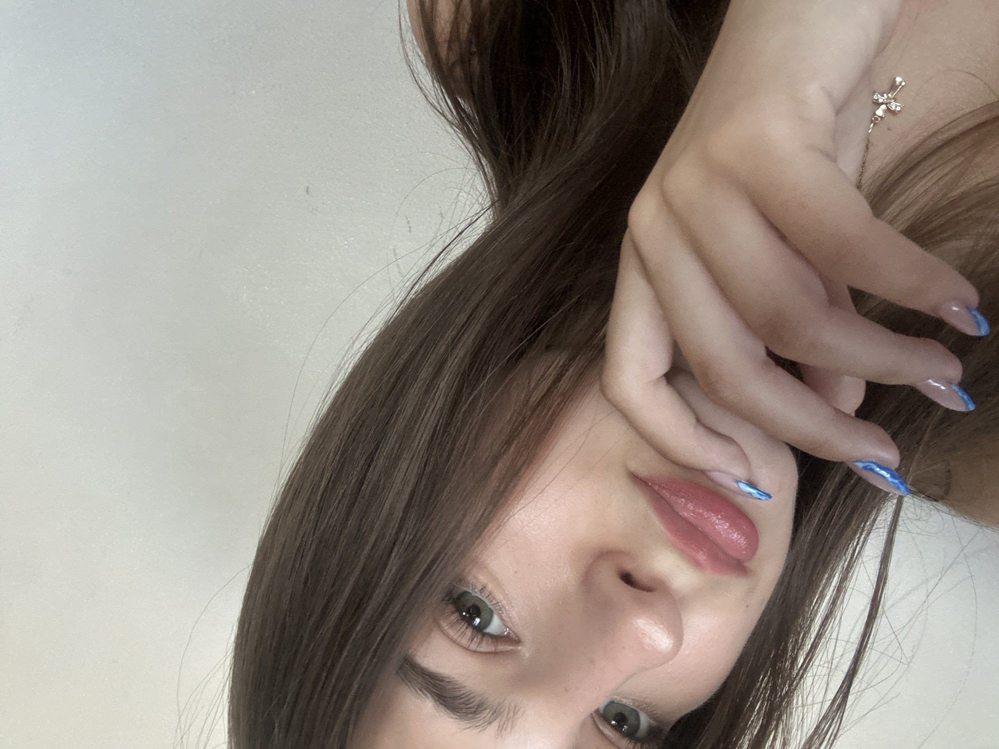 A young woman with long, brown hair is partially visible in the frame. She has green eyes and is wearing pink lipstick. Her right hand is raised to her face, with her fingers touching her chin. She has light blue French manicured nails. A small, gold cross necklace is visible on her neck. The background is a plain, light-colored wall. There is no discernible location in the city of Shumen, Bulgaria.