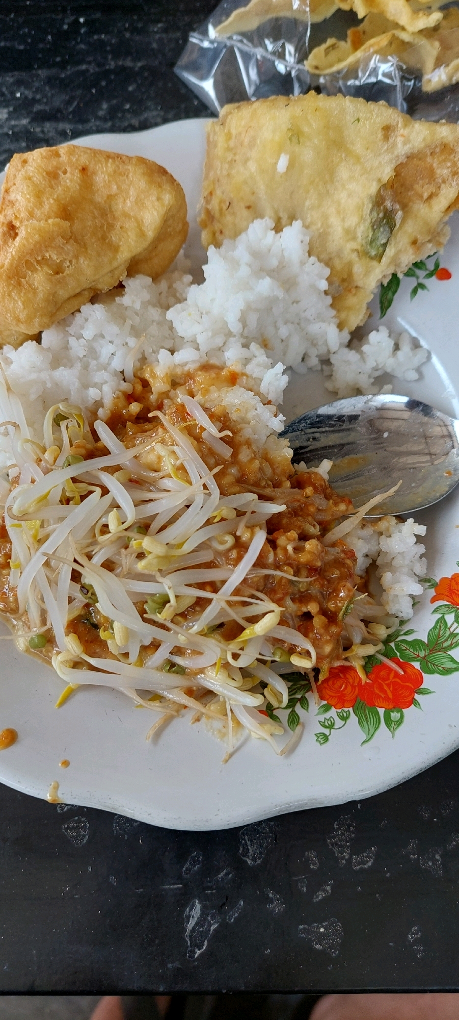 A plate of food is shown from a top-down perspective. The plate is white with a floral design around the rim. The food consists of white rice, topped with bean sprouts and a sauce, possibly peanut sauce. To the left of the rice is a piece of fried food, likely tofu or tempeh. To the right of the rice is another piece of fried food, which appears to be a vegetable fritter. A silver spoon is partially visible on the right side of the plate. In the background, a plastic bag containing some sort of crispy snack can be seen. The plate rests on a dark, textured surface, possibly a table.