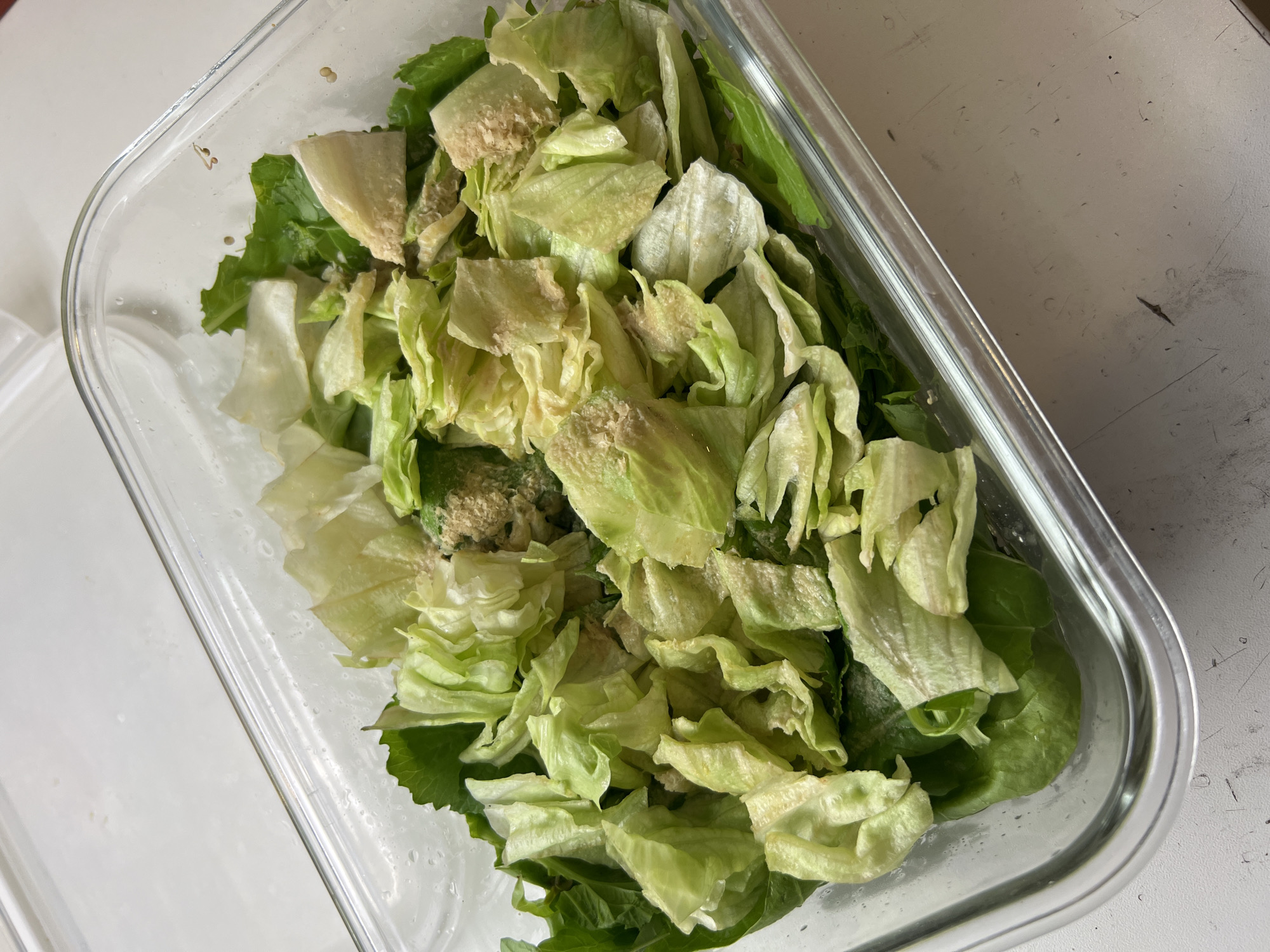 A top-down, close-up shot shows a clear rectangular glass container filled with a mix of lettuce types. The lettuce appears to be a combination of iceberg lettuce, evident from the pale, rounded leaves, and possibly some spinach or romaine, indicated by the darker green leaves at the bottom. Some of the lettuce shows signs of oxidation or browning. Small, unidentified brown specks are also scattered throughout the lettuce. The container appears to be resting on a white surface with some markings or imperfections. The setting and specific location within the city of Unknown, Unknown, cannot be determined from this image alone.