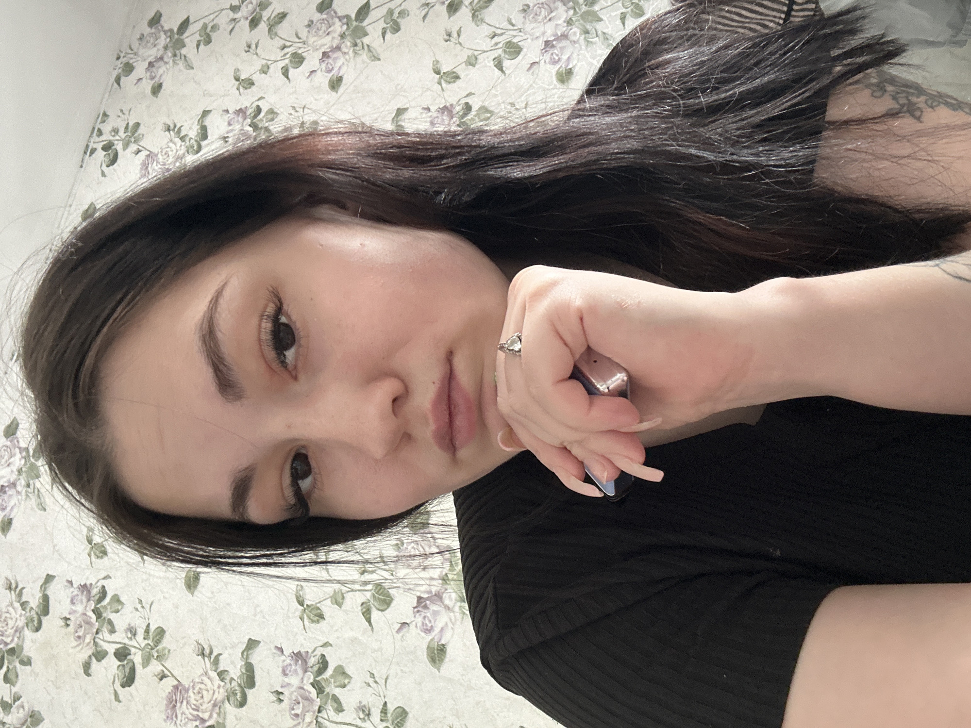 A female individual is depicted in a close-up, selfie-style indoor portrait, identified as being in the city of Chaikovsky, Russia. The subject, an adult female with long, dark brown hair and a fair complexion, occupies the majority of the frame, looking directly towards the viewer with a neutral, slightly pensive expression and mildly pursed lips. She is wearing a black, ribbed, short-sleeved top. Her left hand is raised, with fingers partially curled, resting near her chin. A silver-toned ring with a clear or light-colored stone is visible on her left ring finger. Her long fingernails display a two-tone manicure, featuring a light, possibly natural, base color with darker tips. She holds a small, metallic, silver-colored object, appearing to be a lighter, loosely between the fingers of her left hand. A dark-inked tattoo, possibly floral or abstract, is partially visible on her upper left arm/shoulder area, obscured by hair and clothing. The background consists of a wall covered with light-colored wallpaper featuring a repeating pattern of purple roses and green leaves.