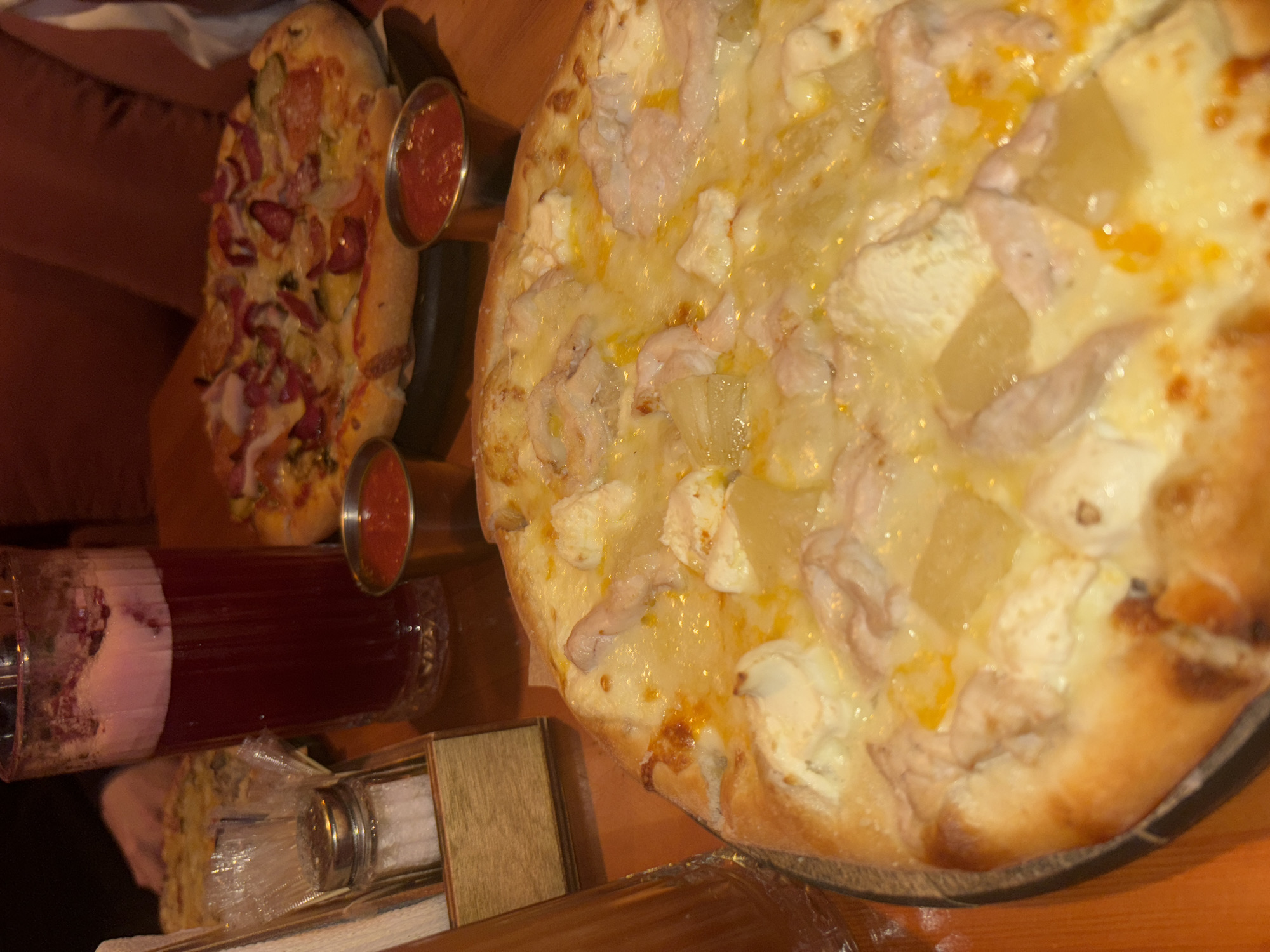 A high-angle shot reveals two pizzas, possibly in a restaurant in Perm, Russia. The pizza on the right is round with toppings that appear to be chicken and pineapple. The pizza on the left is oblong, with sausage, red onion, and possibly green pepper. Two small metal containers of red sauce sit between the pizzas. A long glass with a red liquid (perhaps juice or sangria) and white foam is visible below the left pizza. A wooden box and salt shaker are near the glass. The pizzas rest on a wooden surface with a dark reddish background.