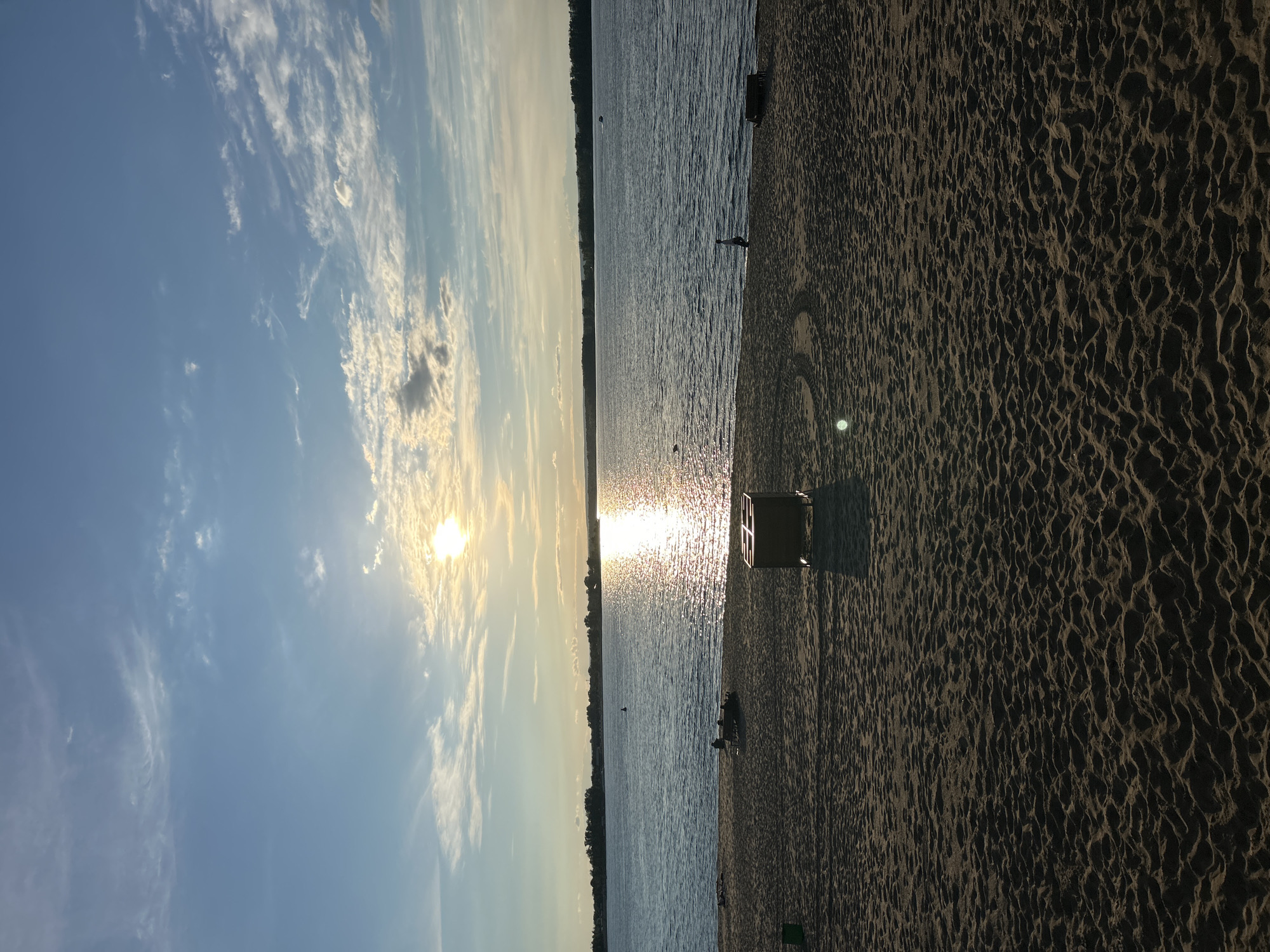 The image depicts a serene beach scene during what appears to be sunset or sunrise. The sun is low in the sky, casting a bright reflection on the calm water. The sky is a gradient of blue with scattered white and gray clouds. In the foreground, a sandy beach is visible, textured with footprints and ripples. Several objects are present on the beach, including a dark-colored trash receptacle, a small wooden bench or table with people seated around it, and a green trash bin. In the water, a person can be seen wading or swimming. The horizon shows a distant treeline and possibly a few structures. The overall impression is one of peacefulness and natural beauty. This scene could be found at the shores of the Volga River, which flows near Novokuybyshevsk, Russia.