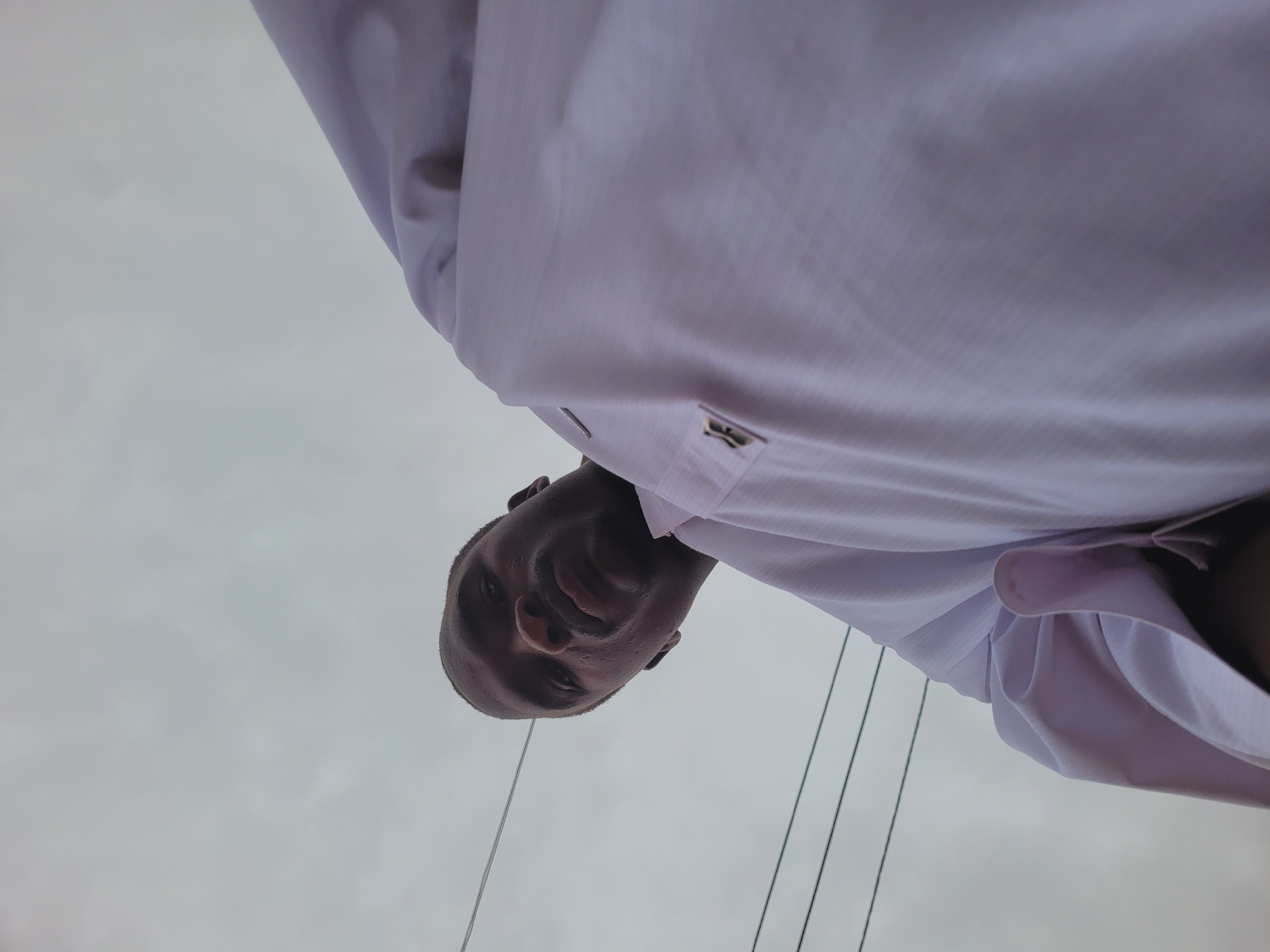 A low-angle shot depicts a man wearing a white collared shirt against a gray sky. The man's face is prominently visible, positioned above the shirt. Thin, dark lines, possibly wires or ropes, extend from the top of the frame down towards the sky. No specific details indicate the location as Bukuru, Nigeria.