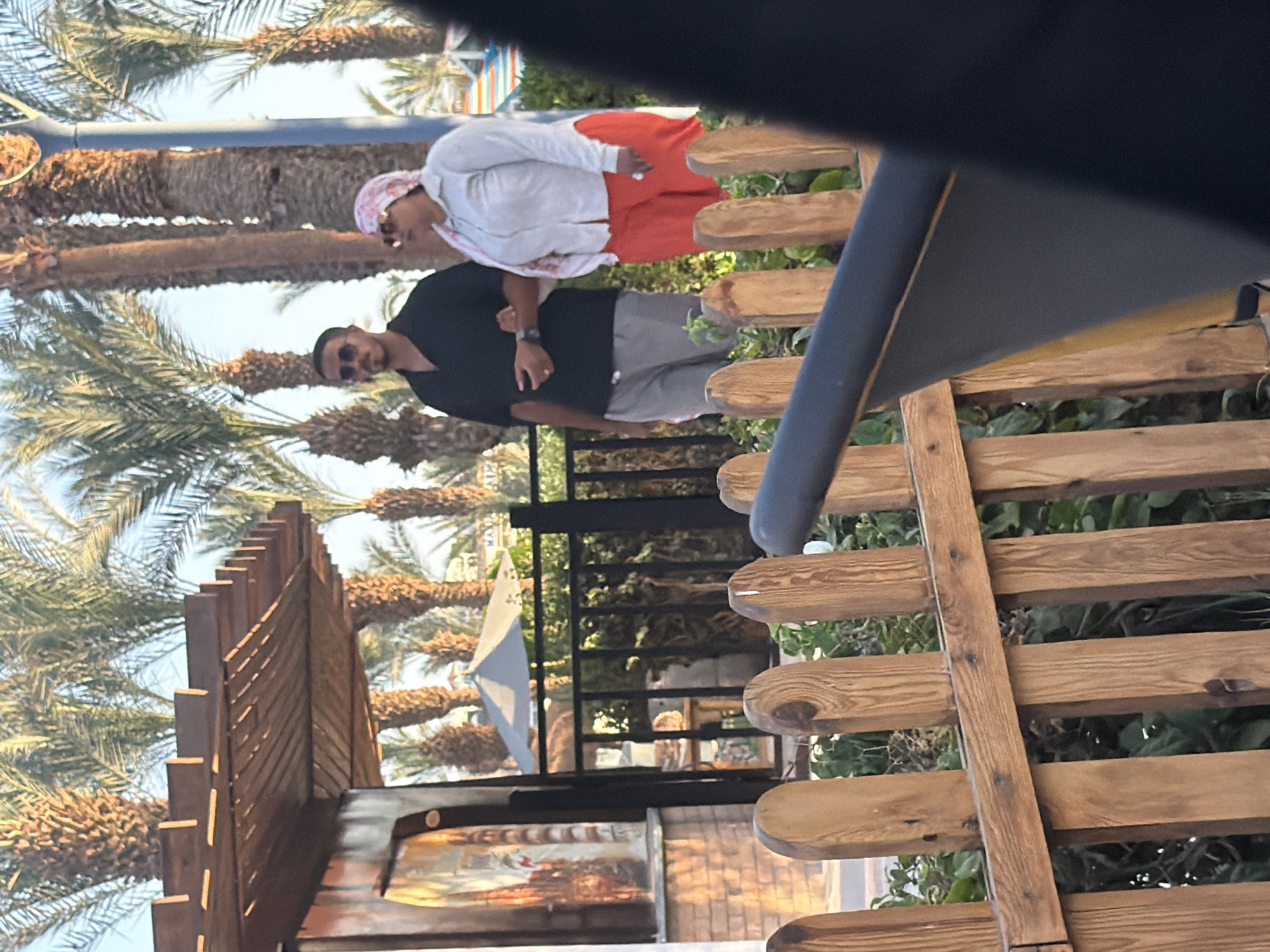 The image captures a street scene in Sharm El Sheikh, Egypt, under a clear sky with abundant palm trees. In the foreground, a low wooden fence is visible, with lush green foliage behind it. To the left, a building with a rustic wooden roof structure and an arched window with a painted mural can be seen. A black metal fence runs horizontally across the middle ground.
Prominently featured in the mid-ground are two individuals. A man, wearing a dark short-sleeved shirt and light gray shorts, stands with his left arm linked through the right arm of a woman. The woman is dressed in a light-colored long-sleeved shirt and bright orange pants, with a pink and white patterned headscarf and sunglasses. They appear to be walking or posing for a photograph.
Behind the couple, several tall palm trees dominate the background, their fronds reaching towards the sky. A light-colored umbrella is visible behind the metal fence, suggesting an outdoor seating area or a recreational space. The overall environment appears to be a warm, possibly resort-like setting, consistent with locations found in Sharm El Sheikh. The lighting suggests it is daytime, likely late afternoon given the angle of the shadows.