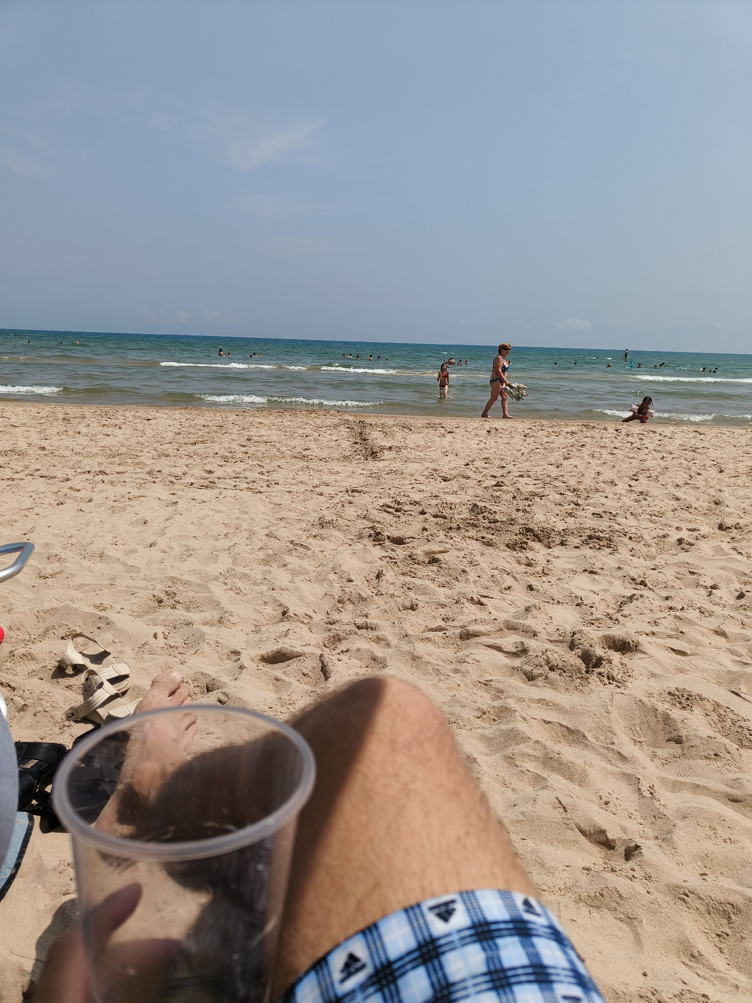 The image depicts a beach scene with a clear sky and calm ocean waters. The foreground shows a person's legs and a plastic cup, suggesting they are relaxing on the sand. Scattered across the sand are a pair of sandals. In the background, several people are visible in the water, some swimming and others standing. A woman in a bikini walks along the shoreline. The beach is made of light brown sand, and the water is a deep blue. The overall impression is one of a pleasant day at the beach. While the image is of a beach, there is no specific visual information to confirm its location in Gandia, Spain.
