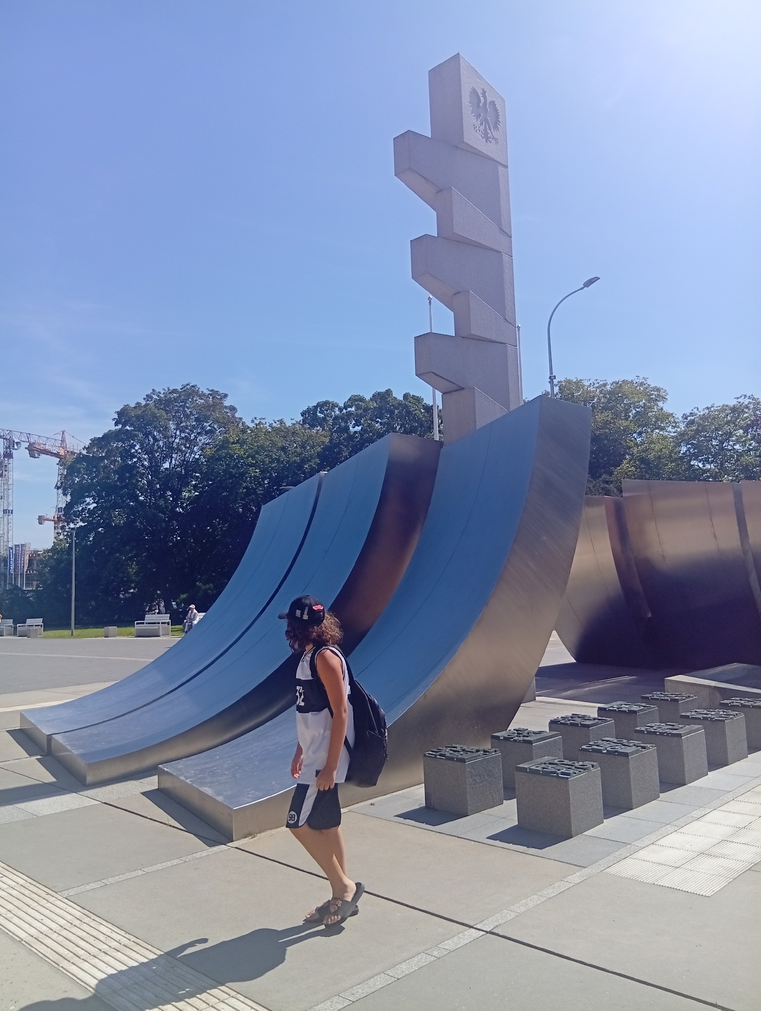 The image shows a person walking in front of a large, modern sculpture in Gdynia, Poland. The sculpture is made of concrete and metal, with curved blue sections and a tall, stepped concrete structure topped with a symbol resembling an eagle. In the background, there are trees, a streetlamp, and construction cranes. The ground is paved with concrete slabs and has a textured strip for tactile paving. The person is wearing a black baseball cap, a white tank top with black trim, black shorts with white accents, and dark sandals. They have a black backpack on and are casting a shadow on the ground. The sky is clear and blue.