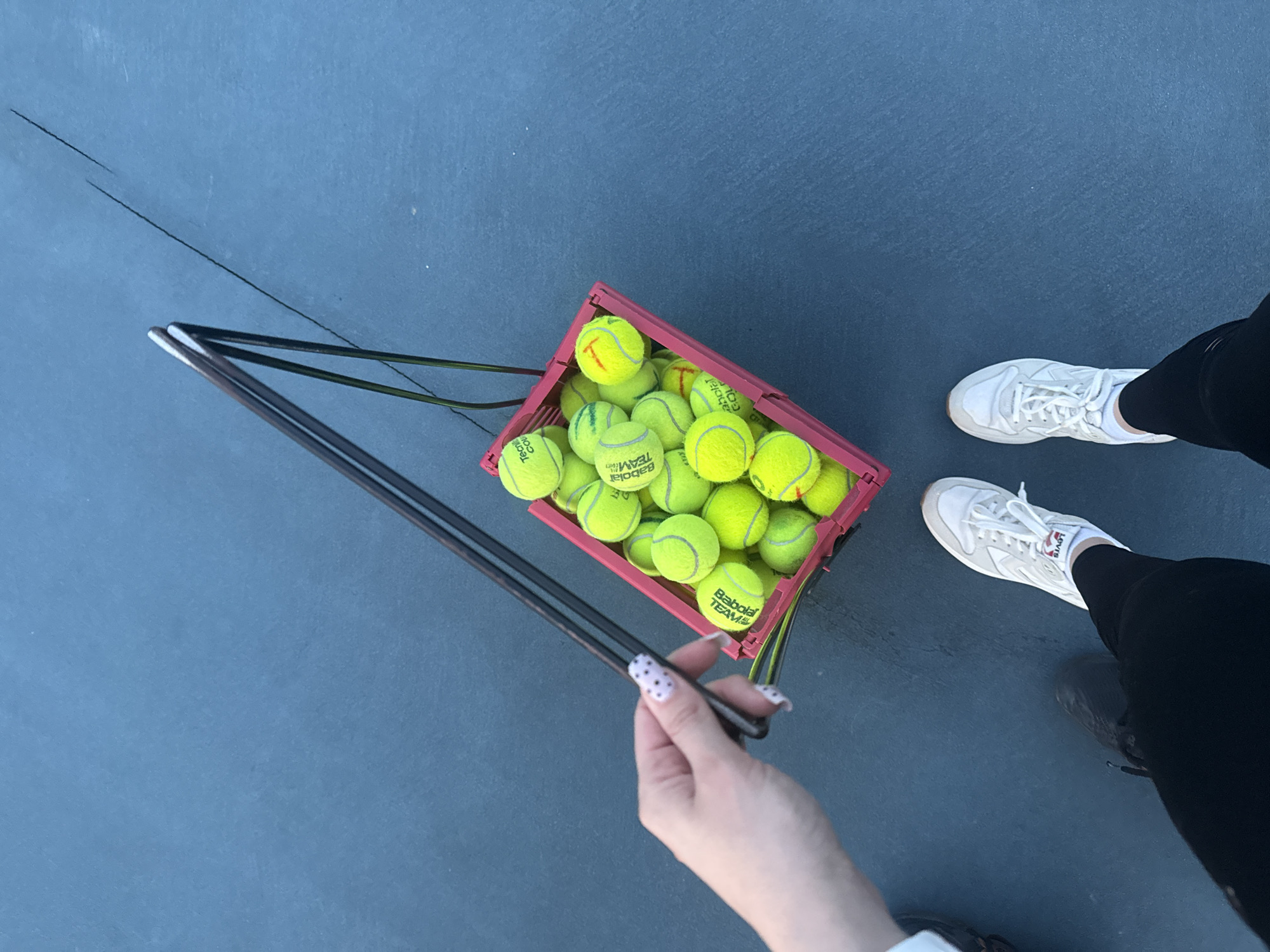 A high-angle shot depicts a blue tennis court. A red plastic basket, filled with yellow tennis balls labeled "Babolat Team," is positioned towards the center of the frame. A black metal tool, possibly used for collecting tennis balls, is held by a hand with polka-dot painted nails. The tool points towards the basket.
To the right of the basket, a pair of legs clad in black leggings is visible, and white sneakers are on the feet. A dark line traverses the blue court surface from the upper left towards the center of the frame. The lighting suggests an outdoor setting, potentially at a tennis facility.
The image does not provide enough information to determine whether the photograph was taken in the city of Batumi, Georgia.