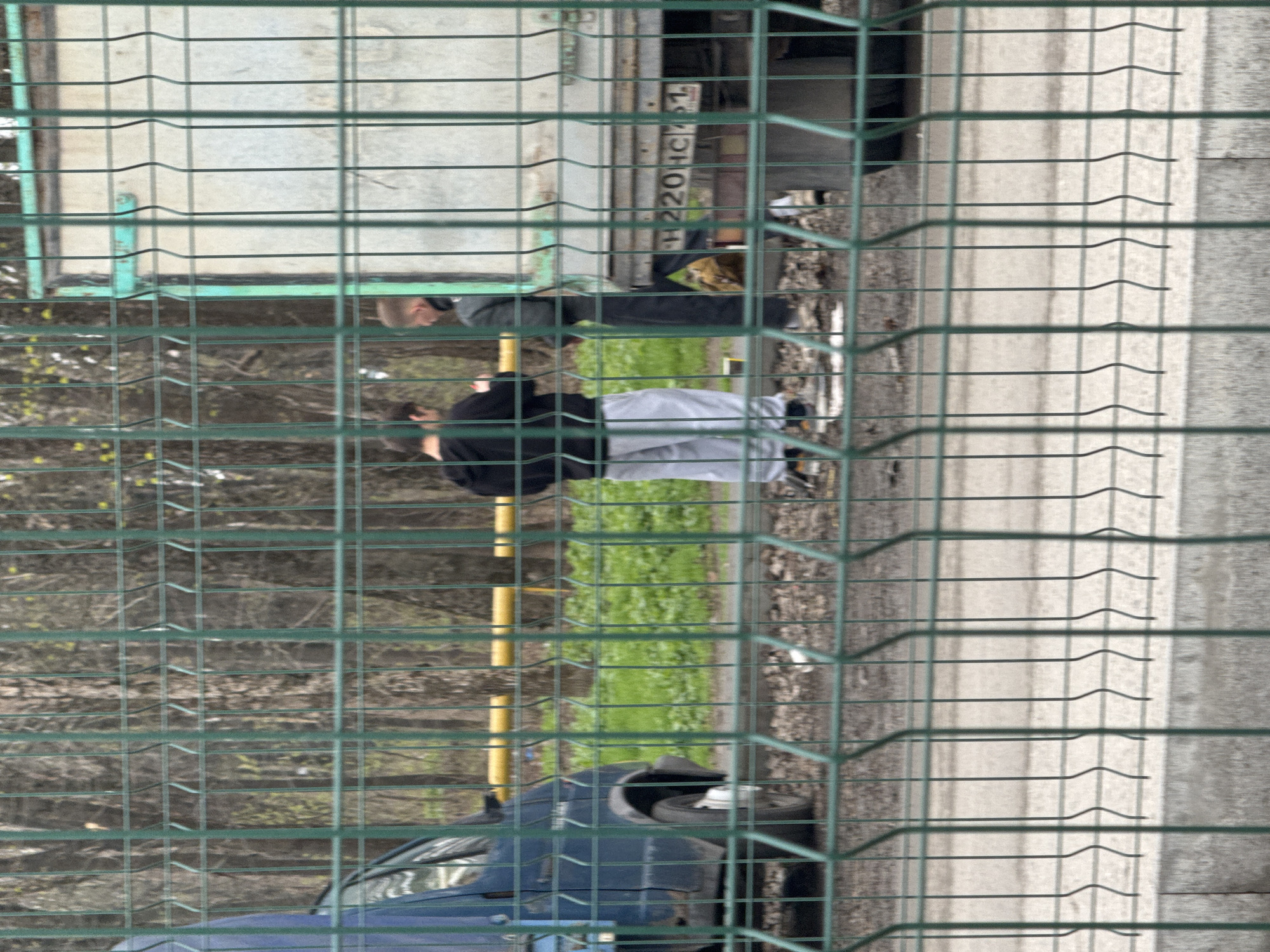 The image shows a fenced area in Rostov-on-Don, Russia. Two individuals are standing near a truck with the license plate "H220HC 61." One individual is wearing dark clothing and light-colored pants, while the other is wearing dark clothing. A blue car is parked near the fence. The environment includes trees, grass, and a paved area.