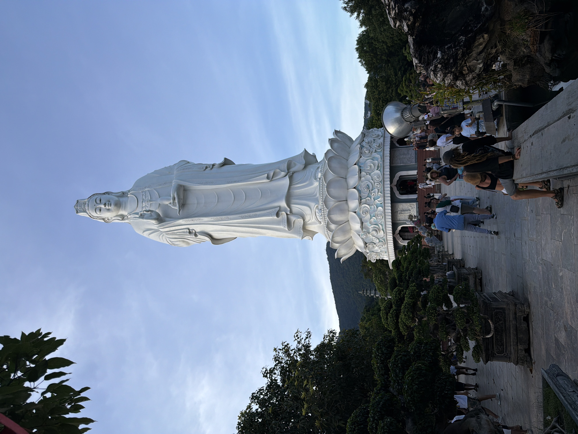 A colossal white statue of a goddess, likely Guan Yin, dominates the frame, standing tall on a multi-tiered lotus-shaped pedestal. The statue, facing forward with a serene expression, holds an object in one hand and has the other raised in a gesture of blessing. The pedestal is intricately carved with floral motifs in blue and white.
Surrounding the base of the statue, a paved walkway leads to a building with an ornate entrance. Lush green trees and well-maintained bonsai trees in stone planters line the walkway. In the background, a conical pagoda and rolling green hills under a bright blue sky with scattered clouds are visible.
A crowd of people, dressed in casual attire, are scattered throughout the scene, walking along the pathways, observing the statue, or gathered near the building. Some are seen from behind, while others face the camera. The overall impression is a popular tourist attraction, likely part of a religious complex, in Đà Nẵng, Vietnam.