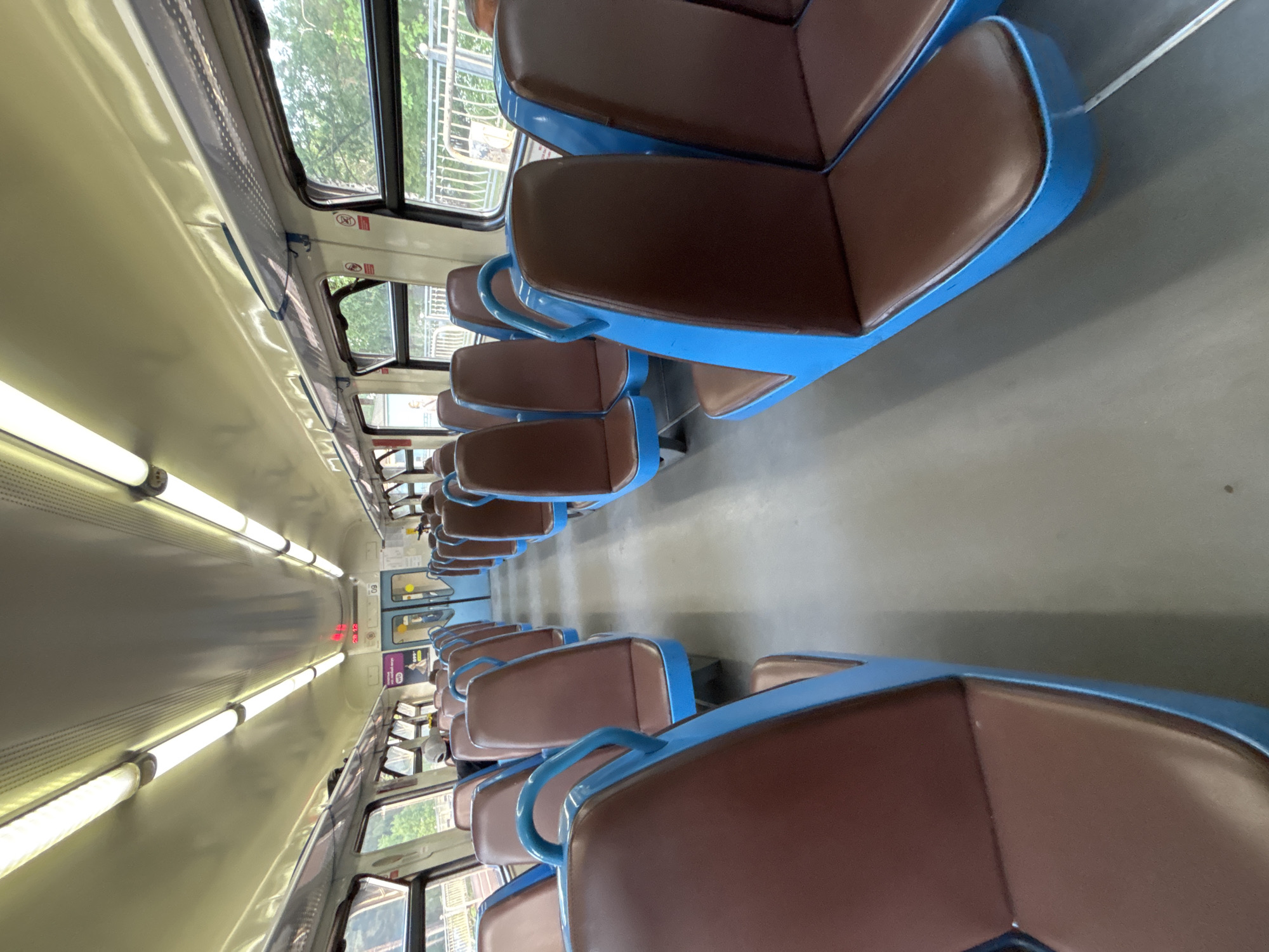 The image shows the interior of a train carriage, likely a commuter train or "elektrichka," consistent with the Komarovo, Russia location context. The perspective is tilted 90 degrees, looking down a long, central aisle between two rows of seats.
The seating features warm brown upholstered cushions set within distinct bright blue frames and armrests. The seats are arranged in pairs, facing each other across the aisle. The floor of the carriage is a utilitarian gray, contrasting with the light beige or off-white ceiling, which is fitted with long, bright fluorescent or LED tube lights running the length of the carriage.
Large windows line both sides, revealing green trees and some distant structures, indicating daylight and a journey through a natural or suburban setting. Blue grab handles or horizontal bars are visible above the windows on the left side, likely for standing passengers.
Towards the far end of the carriage on the left, a single passenger is partially visible, seated. In the distance, a red digital display shows the numbers "20 12," which could indicate the time or a route number. Above this display, the number "60" is faintly visible. Small, red pictogram signs are also present above the windows on the right side. The carriage appears clean and mostly empty, suggesting it is not at peak occupancy.