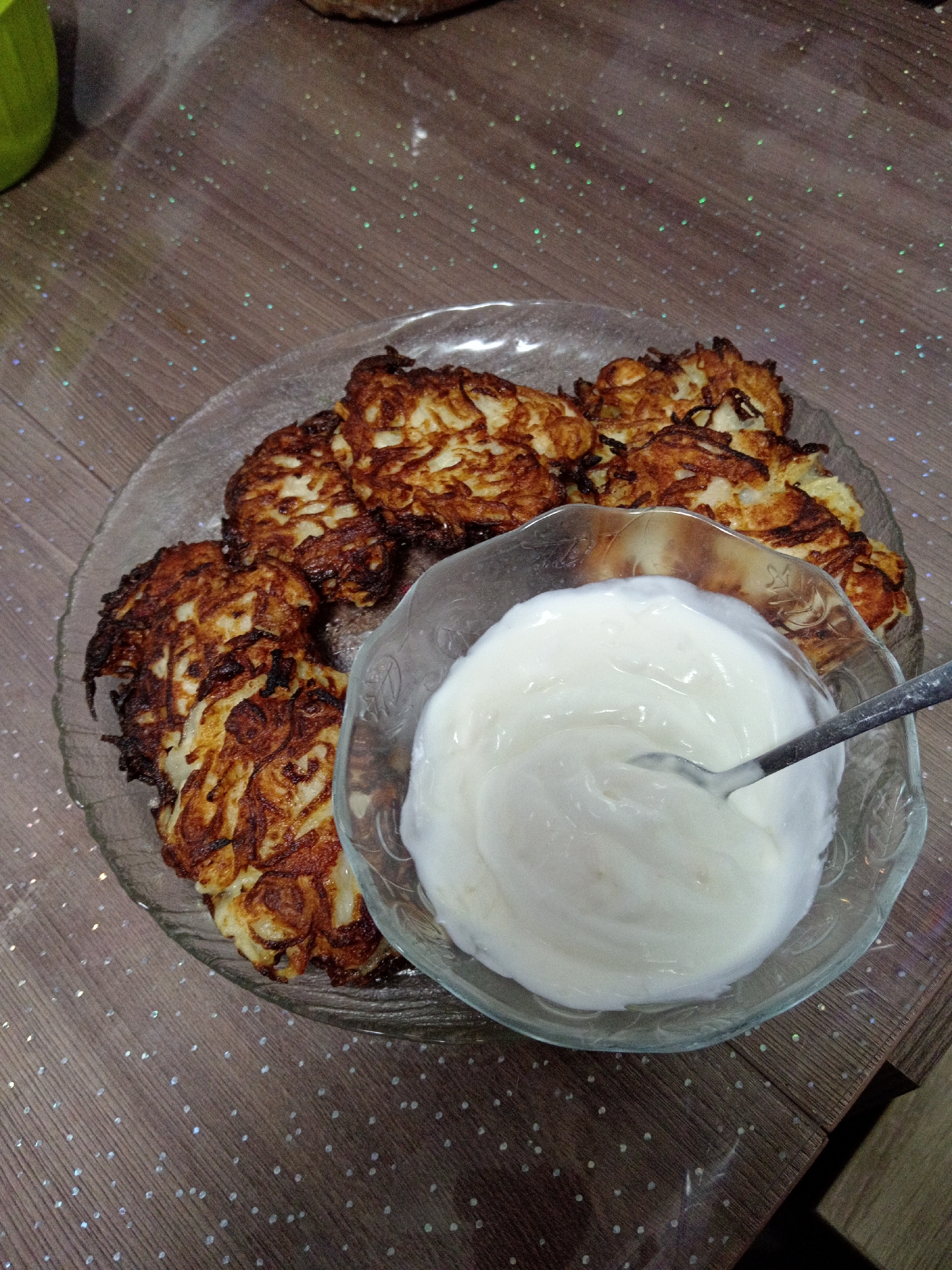 The image shows a close-up, overhead view of a plate of potato pancakes served with a bowl of sour cream. The potato pancakes are golden brown and appear to be crispy. The sour cream is white and creamy, and a spoon is resting in it. The food is placed on a wooden table with a speckled, glittery surface. The scene is indoors, likely in a home or restaurant setting. There are no people visible in the image. The location is not specified in Belogorsk, Russia.