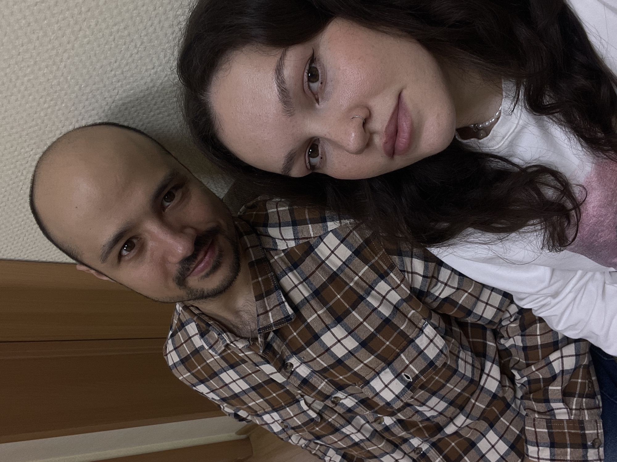 This is a close-up, slightly rotated selfie of two individuals, a man and a woman, posing together indoors in Izhevsk, Russia. The shot is angled upwards, with the woman resting her head on the man.
The man, on the left, has a shaved head or receding hairline and a dark beard and mustache. He wears a brown, white, and dark blue plaid button-up shirt with several buttons visible down the front and on the cuffs. He looks directly at the camera with a calm, slightly smiling expression.
The woman, on the right, has long, dark, wavy hair that frames her face and spreads around her. She has brown eyes, styled eyebrows, and is wearing eyeliner and lip color. A small silver ring is visible in her left nostril. She is wearing a white top, and a pearl necklace with a small charm adorns her neck. A light pink or purple fabric is visible behind her, possibly part of her attire or a blanket. Her gaze is directed towards the camera with a neutral, relaxed expression.
The background behind the man shows a light-colored, textured wall, possibly wallpaper, and a section of brown wooden panelling. The setting appears to be a comfortable indoor space. No text is visible in the image.