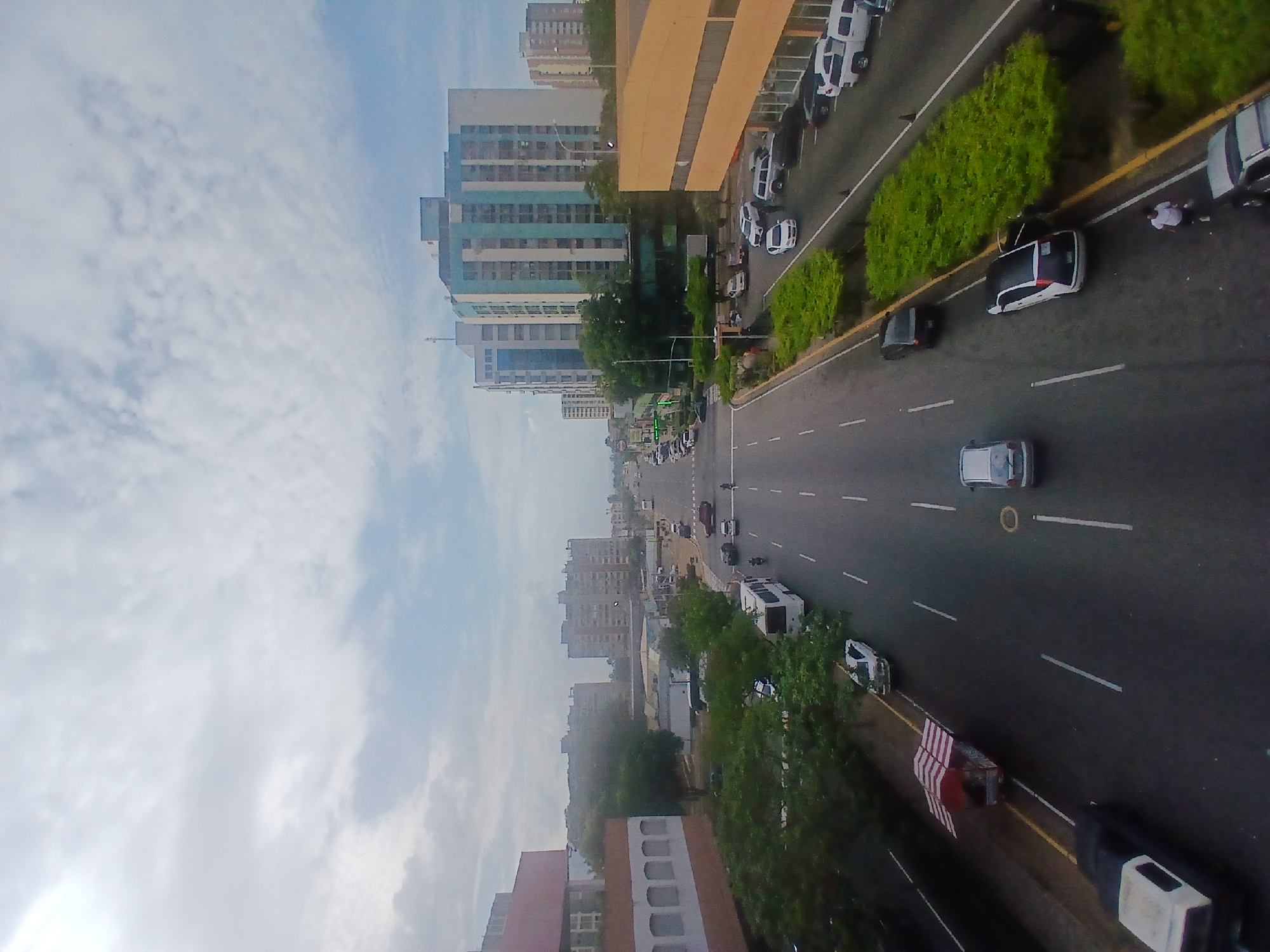 An elevated view captures a vibrant urban street scene in Ciudad Guayana, Venezuela, under a partly cloudy sky during daylight hours. The image is taken from a high vantage point, looking down onto a multi-lane road flanked by buildings and green spaces.
Numerous vehicles populate the scene. Cars, including sedans, SUVs, and a white bus, are seen driving on the asphalt road, which features white lane markings and a yellow curb line. Many vehicles are parked along the side of the road, particularly on the right. One white car on the right side of the road has its trunk open, with a person in a white shirt standing nearby, possibly attending to the vehicle or loading/unloading items. Another car, a dark-colored sedan or SUV, is parked further down the road, its door slightly ajar.
Multi-story buildings dominate the background and sides of the road. To the left, several residential or commercial buildings rise, one notable for its blue-green sections and numerous windows. On the right, a large building with a distinctive yellow-beige upper facade and a glass-fronted lower section stands prominently. Lush green bushes and trees line the road's median and sidewalks, adding greenery to the urban landscape. A red and white striped barrier is visible on the median, and a yellow circular marking is on the road. No discernible text is visible.