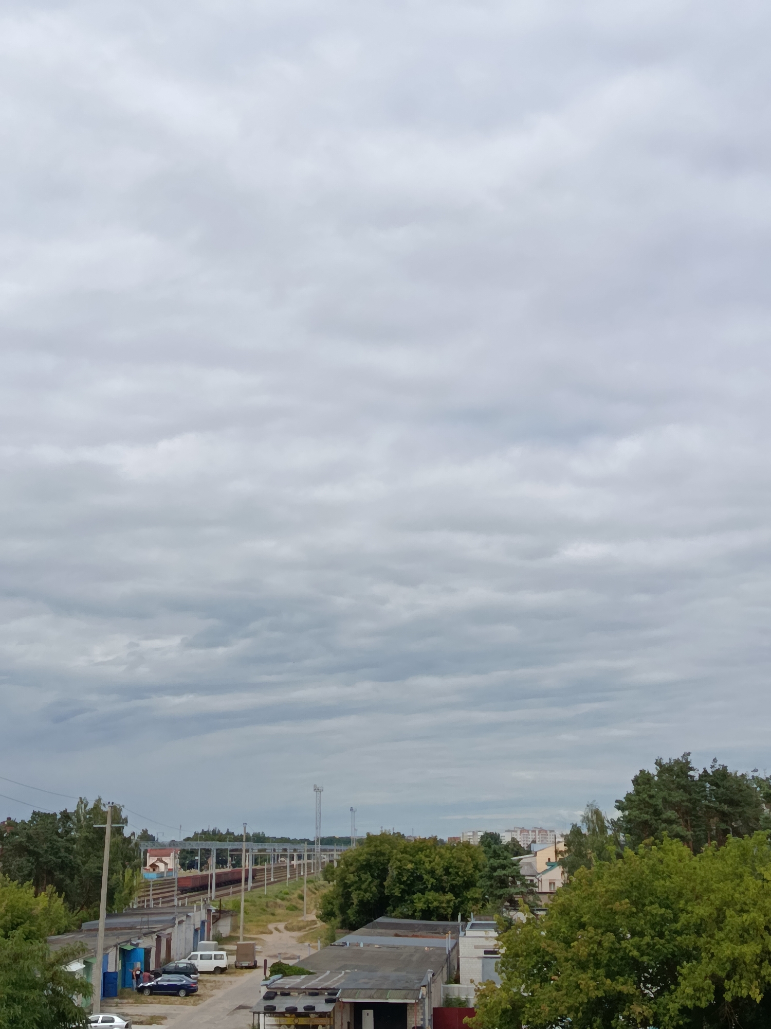 The image captures a wide view of a cloudy sky over a suburban area, possibly in Svietlahorsk, Belarus. In the foreground, a row of garages with corrugated metal roofs are visible. A street runs alongside these garages, with several cars parked, including a blue sedan and a white van. A few individuals are standing near one of the garage doors. Further in the distance, a train with red carriages is traveling on elevated tracks. Beyond the train tracks, a cluster of trees and buildings, including apartment blocks, are seen. The overall impression is a typical overcast day in a residential or industrial fringe area.