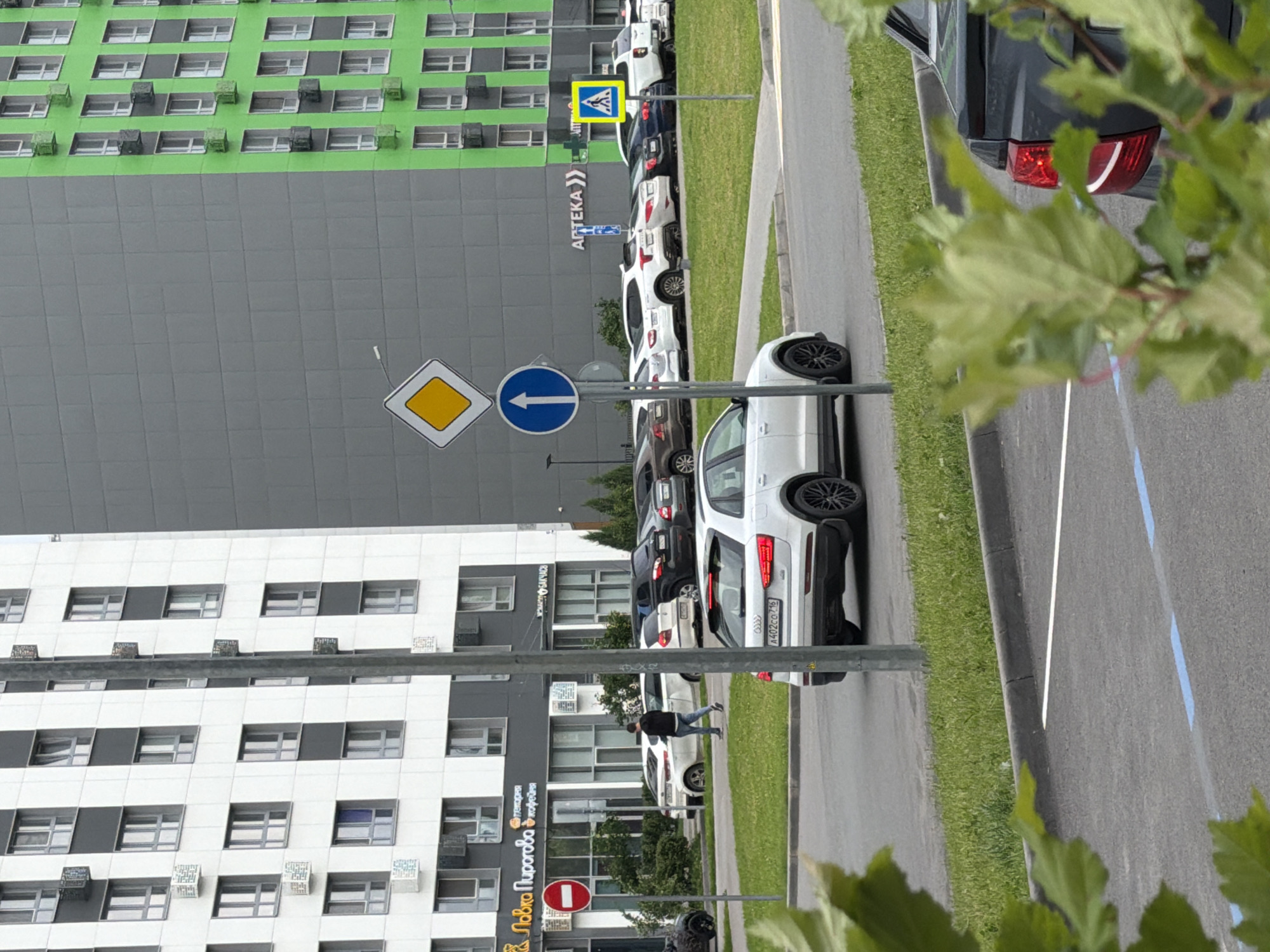 The image depicts a street scene in Kazan, Russia, likely during the daytime on a cloudy day. In the foreground, green leaves partially obscure the view of parked cars along a road. Several white and dark-colored cars are parked in a row, with one white car in the foreground showing its rear and side.
Behind the cars, a modern building complex stands, characterized by its green and grey facade. Numerous windows are visible on the buildings, with some appearing to have air conditioning units attached. A pedestrian crossing sign with a blue circular background and a white arrow pointing left is prominent on a pole, alongside a diamond-shaped road sign with a yellow center. Another sign with a red circle and a white horizontal bar, indicating "no entry," is also visible.
Further down the street, a building with "АПТЕКА" (Apteka, meaning Pharmacy) written on its facade can be seen, along with other signage in Russian, including "ЛАВКА Пирогова" (Lavka Pirogova, a bakery). A person in a dark jacket and blue jeans is walking on the sidewalk. The overall scene suggests a typical urban environment with shops, residential buildings, and a parking area.
