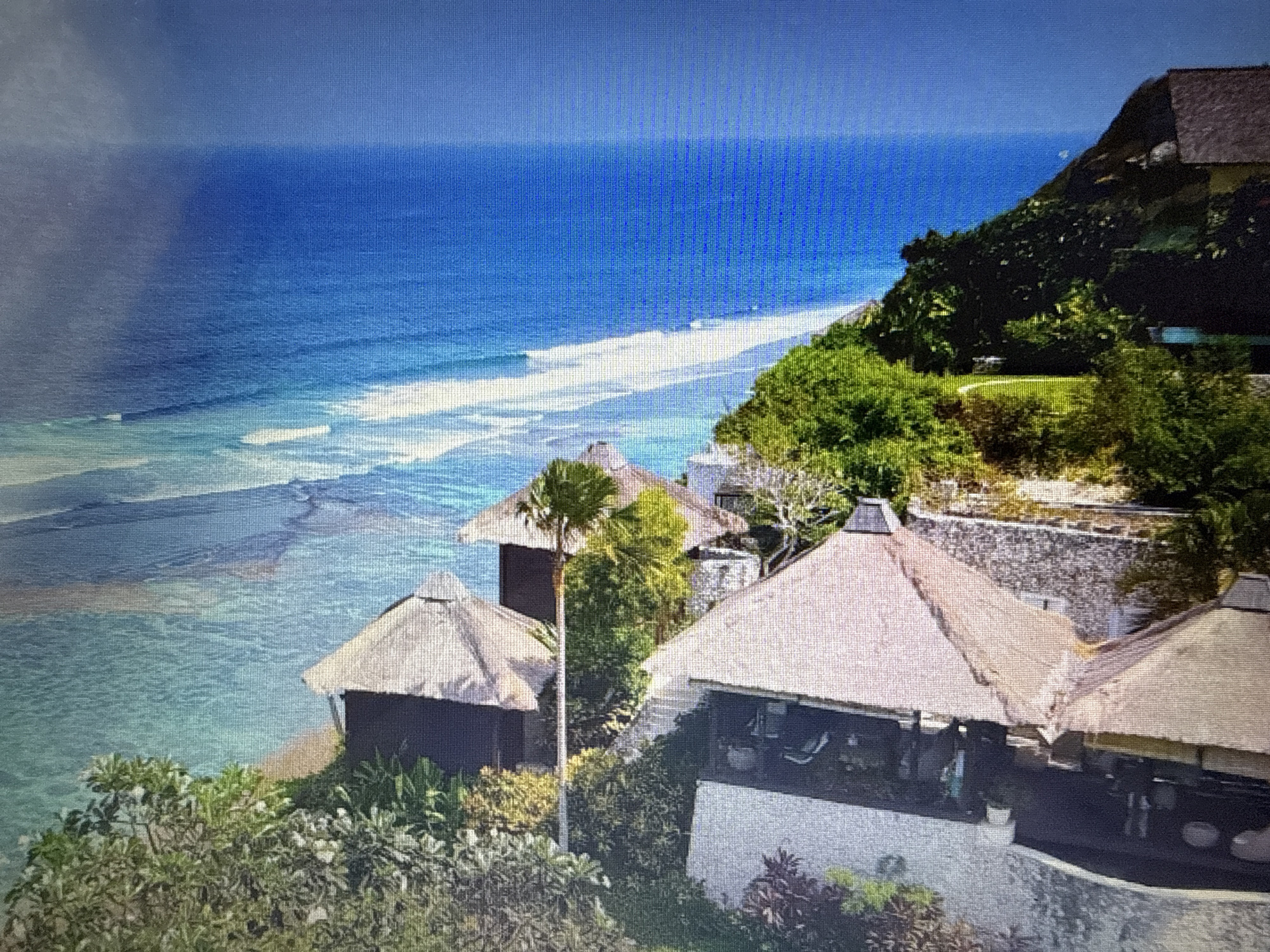 This image depicts a scenic coastal villa with a view of the ocean. The villa features traditional thatched-roof structures and is nestled amidst lush greenery on a hillside overlooking the sea. The ocean is a vibrant blue, with white waves breaking near the shore, suggesting a tropical or subtropical climate. The sky is clear, indicating fair weather.
The villa itself appears to be a resort or private residence, with open-air living spaces and lounge chairs visible on a terrace. Several buildings with conical thatched roofs are scattered across the property. In the foreground, dense foliage and palm trees contribute to the natural beauty of the setting.
While the image is primarily a landscape, there are indications of human presence. Silhouettes of people can be seen on the terrace of the main villa building, suggesting the property is occupied. The time of day appears to be daytime, likely late morning or early afternoon, given the bright sunlight and shadows.
There is no visible text in the image. The overall mood conveyed by the image is one of tranquility, luxury, and natural beauty, characteristic of a high-end tropical vacation destination. The location context provided, Pushkino, Russia, seems incongruous with the visual elements of the image, which strongly suggest a warm, coastal environment.