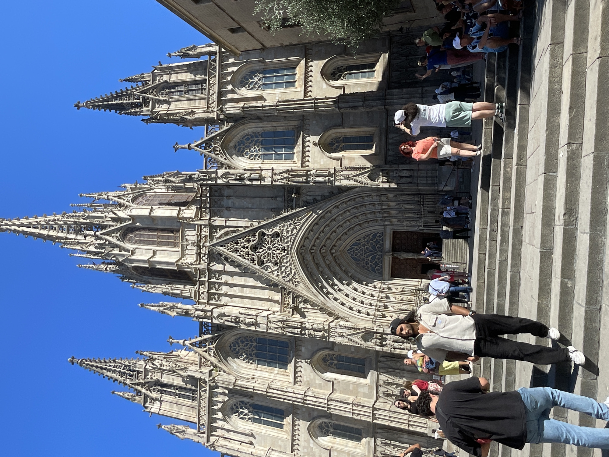 The image shows a view of the Barcelona Cathedral, located in the Gothic Quarter of Barcelona, Spain. The camera angle is low, emphasizing the facade. The cathedral is a Gothic-style building with intricate stonework, pointed arches, and towering spires against a clear, bright blue sky.
The foreground includes a set of stone steps leading up to the cathedral's entrance. Several people are on the steps, including a man in a light-colored shirt and black pants, a person in a black shirt and blue jeans, a woman in an orange top, and another person in a white top. They appear to be tourists or visitors.
The scene is bathed in bright sunlight, creating strong shadows on the building and steps. The image captures a moment in time, with people interacting and posing for pictures in front of the cathedral.