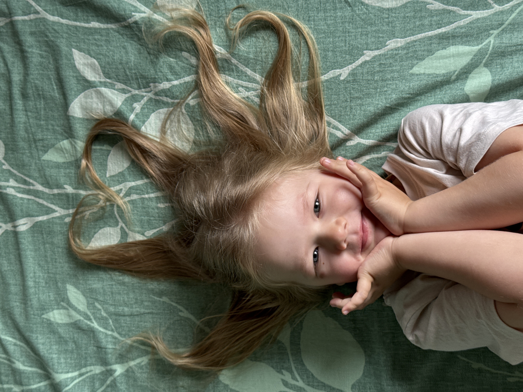 A young girl with blonde hair is lying on her back on a green bedspread with a white leaf pattern. Her hair is spread out around her head. She is wearing a light-colored shirt and has her hands near her face. The location is not discernible from the image.