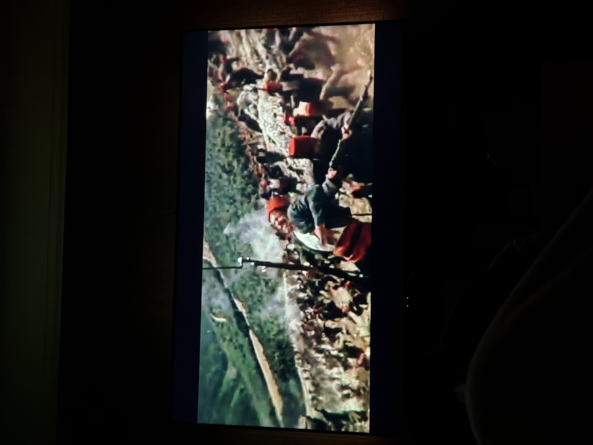 A television screen displays a scene with numerous soldiers, some wearing red headgear, engaged in what appears to be a battle. Some soldiers are holding rifles. The background features what looks like terrain with green areas. The lighting conditions are dark in the room where the television is located. The city and state are unknown.