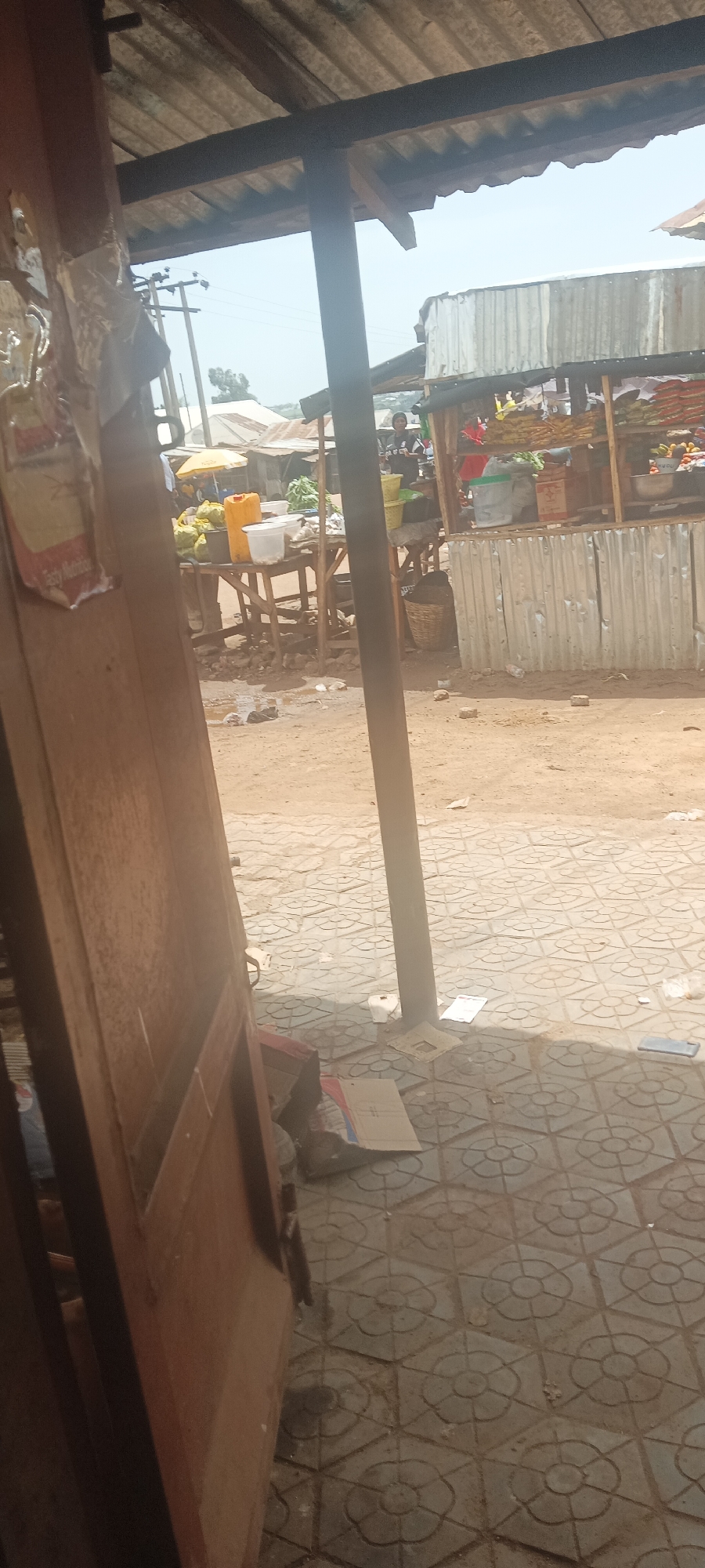 The image depicts a scene in Bokkos, Nigeria, likely a market area. The view is partially obscured by the frame of a building, including a wooden door on the left. The foreground features a tiled floor with a floral pattern, scattered with debris.
Through the building's frame, a bustling marketplace is visible. There are several stalls, one constructed with corrugated metal sheets, displaying a variety of goods, including what appears to be fruits, vegetables, and possibly other food items. People are present in the market, some standing and engaging in what seems to be purchasing or selling activities. There are also containers such as water tanks or buckets, and baskets nearby. The background shows other buildings and the sky. The overall environment appears to be a busy, open-air marketplace.