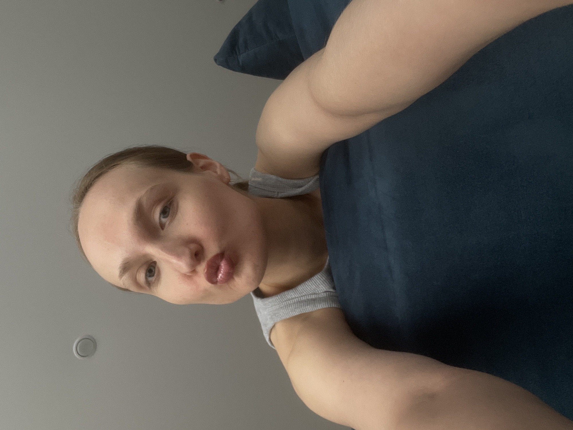 A person is positioned in front of a blue cushion or piece of furniture, likely indoors. The background includes a grey wall with a circular vent or fixture. The person is wearing a grey sleeveless top. The location cannot be confirmed as Podshivalovo, Russia, based on the image.