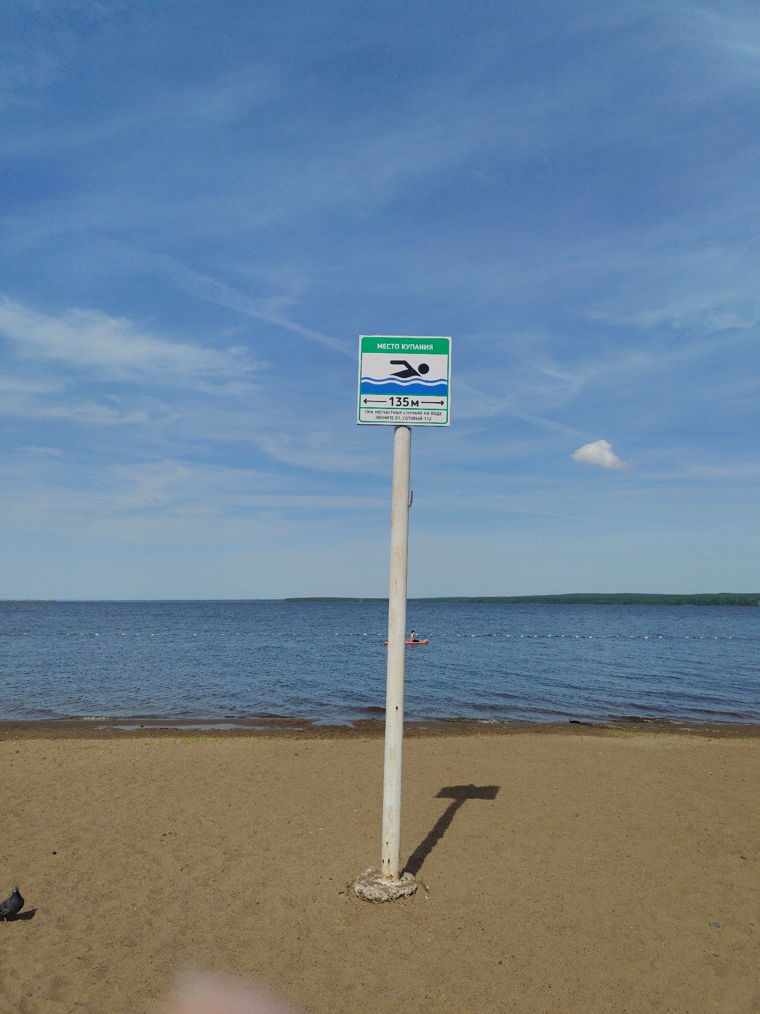 A daytime shot showcases a sandy beach leading to a body of water under a partly cloudy sky. A signpost stands prominently on the beach, displaying a symbol of a swimmer and text indicating "Swimming Area 135m" in Russian. Text also includes instructions to call 01 or 112 (mobile) in case of accidents in the water. A single individual on a paddleboard is visible in the water, marked by a line of buoys. The ground near the post shows its shadow and a nearby pigeon.