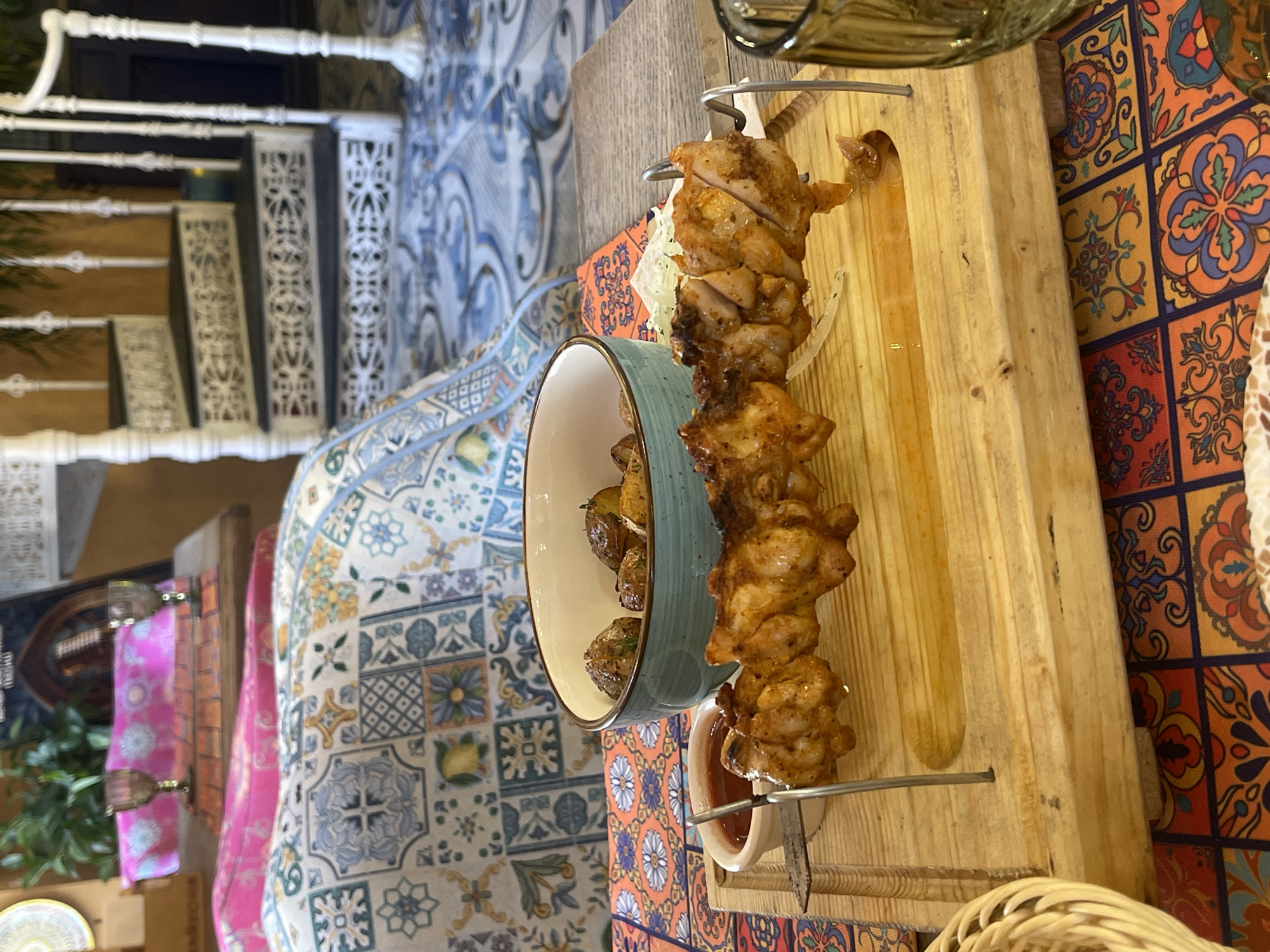 The image shows a table in an establishment, likely a restaurant, that is located in Saint Petersburg, Russia. The table is set with a meal of chicken kebabs served on a wooden platter with a side of roasted potatoes in a light blue bowl. There is also a small bowl containing a dipping sauce. The table is covered with a colorful, patterned tablecloth that seems to be inspired by traditional tile work. The background includes elements of architectural details, possibly inside the establishment itself, like a white, ornate staircase. The environment suggests a space with character, perhaps an eatery that has incorporated traditional or local design elements.