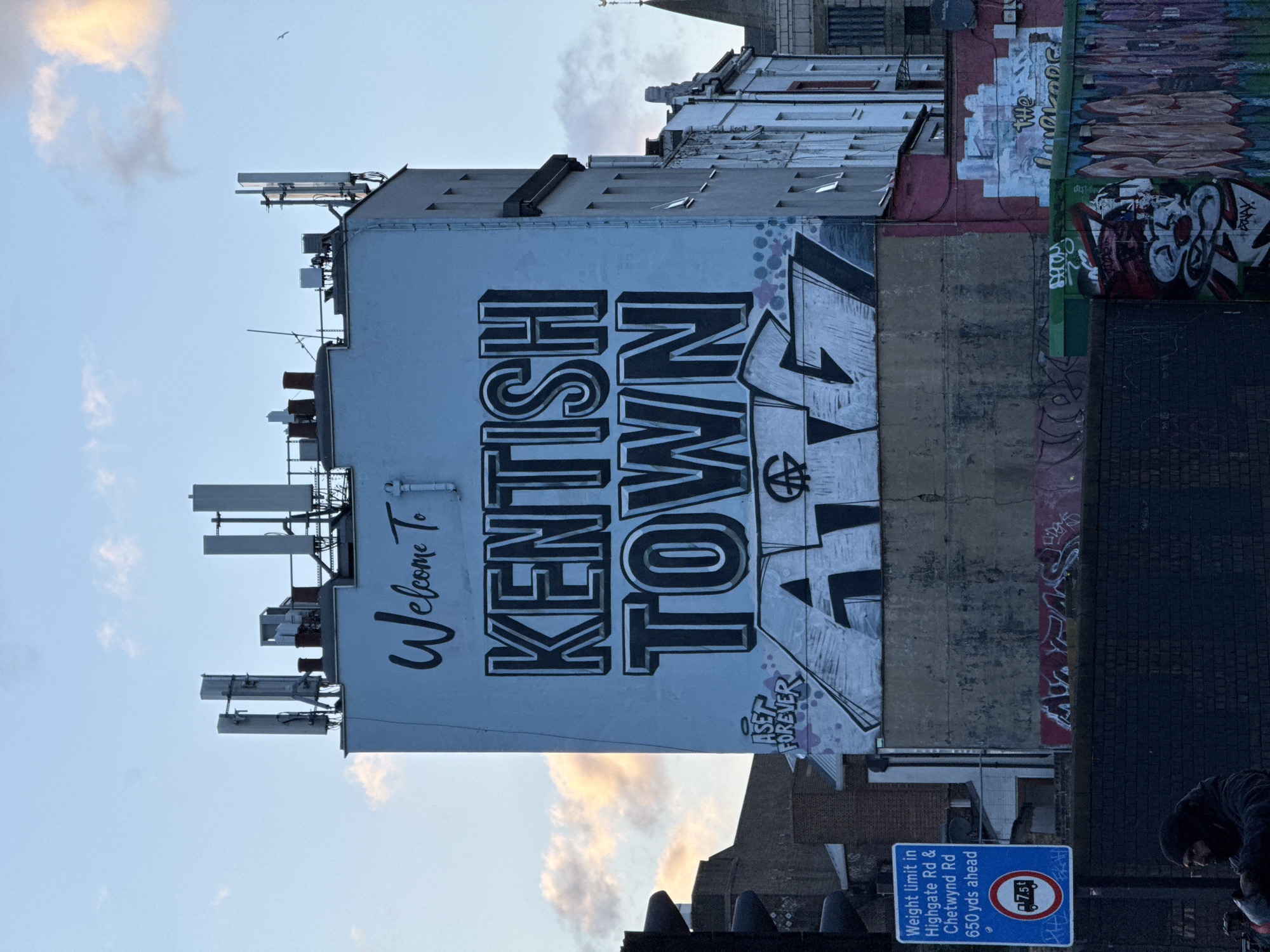 The image shows a building in Kentish Town, London, United Kingdom. The side of the building has a large mural that reads "Welcome To Kentish Town." The mural is painted in a graffiti style with the words "ASET FOREVER" at the bottom. Above the mural are several antennas and other equipment. Adjacent to the painted wall is a section with various forms of graffiti and tags.
In the foreground is a street sign indicating a weight limit on Highgate Road and Chetwynd Road, 650 yards ahead. A person is partially visible in the lower right corner of the frame. The sky is visible with some clouds.