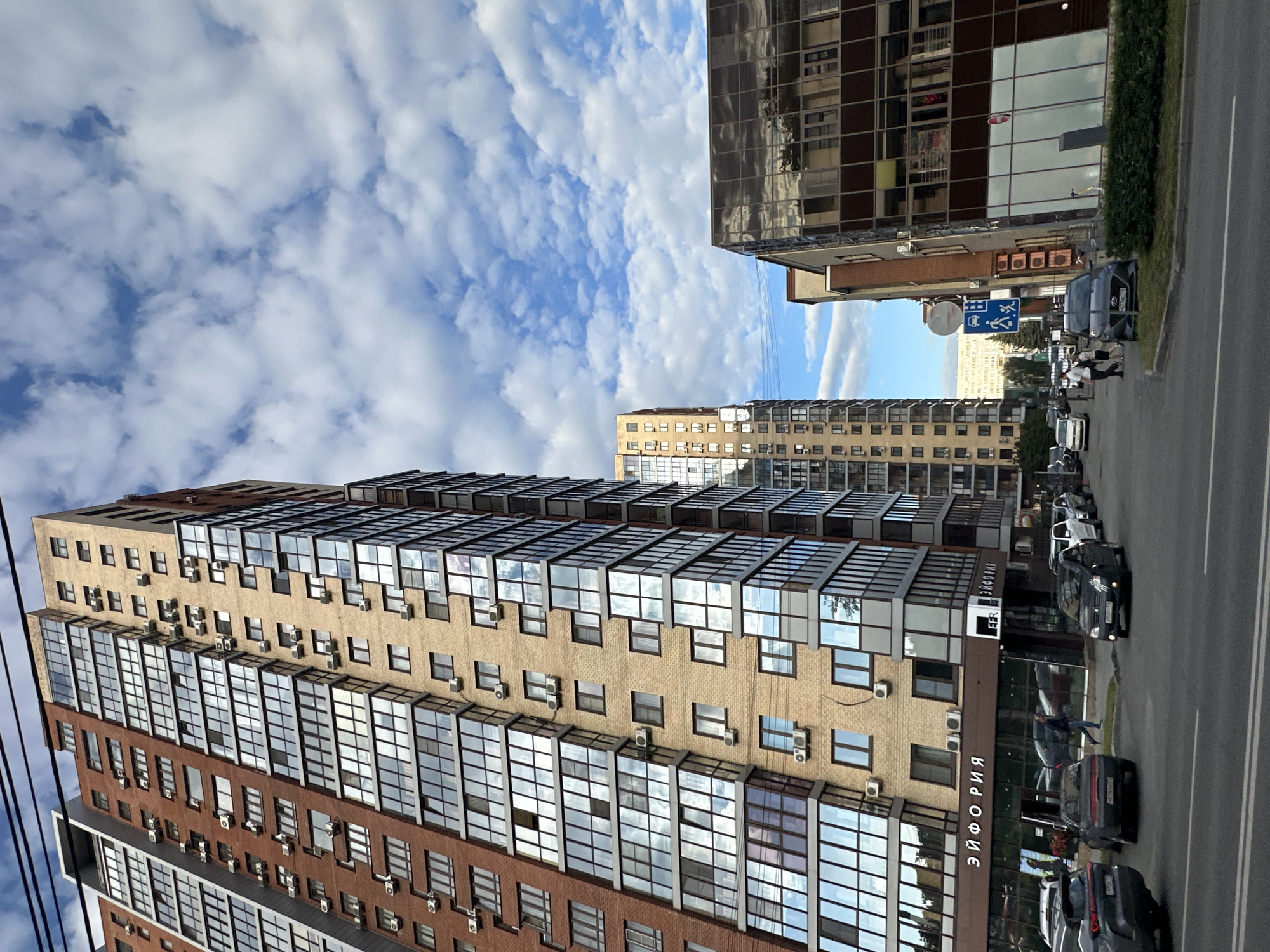 A street scene in an urban environment shows several tall, modern apartment buildings under a partly cloudy sky. On the left, a multi-story brick building with a brown and tan facade features numerous balconies and windows. To its right, a narrower, lighter colored building also has many windows and air conditioning units mounted on its exterior. In the mid-ground, another tall building with a beige facade is visible. Further in the background, more residential structures can be seen. A dark, modern building with reflective glass windows is situated on the right side of the frame, appearing to be a commercial space with an awning above its entrance. A street with several cars parked and driving is in the foreground. A sign with Russian text, "ЭЙФОРИЯ," is visible on the commercial building. Two pedestrians are walking on the sidewalk in front of the commercial building. A blue traffic sign showing a pedestrian crossing symbol and a prohibition symbol is mounted on a pole. The overall impression is a contemporary city neighborhood.