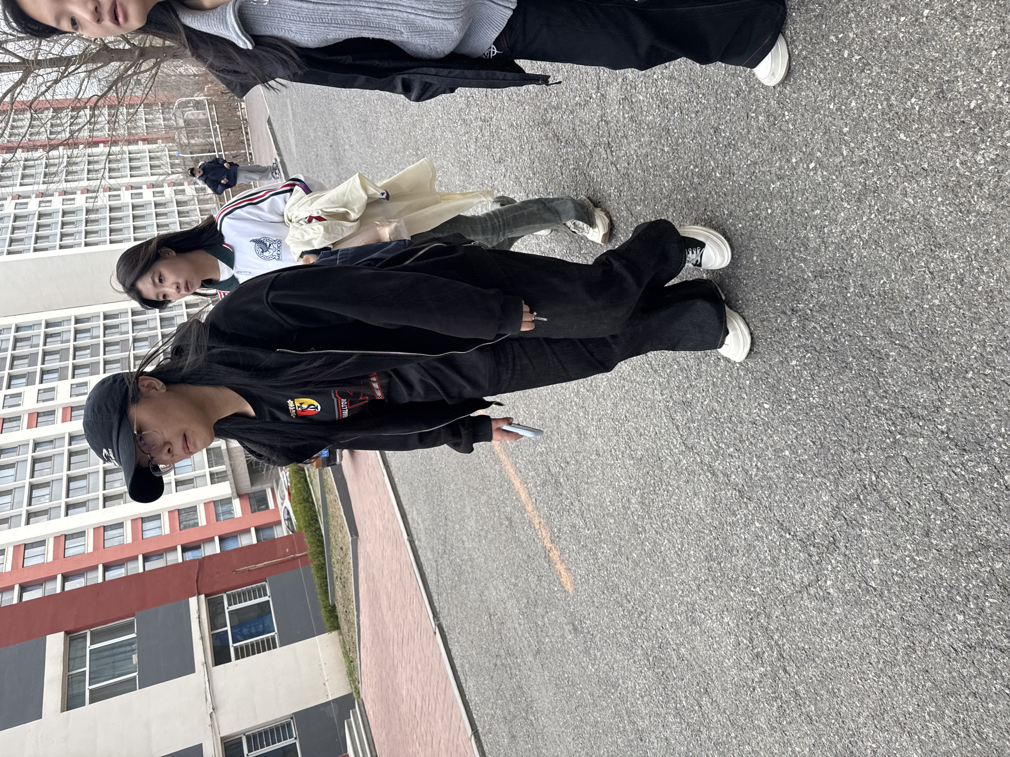 A group of three individuals is seen walking along a paved road in Zhongtun, China, with buildings in the background. The foremost person is wearing a black jacket, pants, and hat, and holding a smartphone. The second person is wearing a white jacket with dark green accents and light blue jeans, holding a tan jacket. The third person is wearing a grey top and dark pants. The setting appears to be an urban environment.