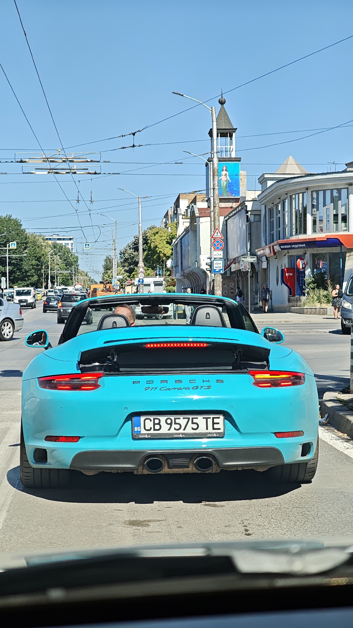 The image captures a bright, sunny day in Sofia, Bulgaria, viewed from inside a vehicle. The primary subject is a vibrant turquoise Porsche 911 Carrera GTS convertible, seen from the rear. The car's license plate reads "CB 9575 TE". The car is in traffic on a city street, indicated by the presence of other vehicles, traffic lights, overhead power lines for public transport, and buildings lining the road.
The urban environment includes a variety of buildings, some with modern architectural elements and others with more traditional facades. A notable feature in the background is a tower with a spire, topped with a decorative element, and adorned with a large advertisement featuring a woman in a mermaid costume. The street is paved, and there are sidewalks with parked cars and pedestrians visible. The sky is clear blue. The scene appears to be a typical urban street in Sofia during daylight hours.