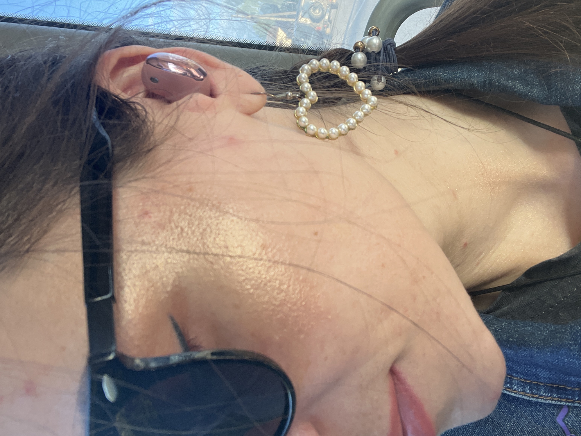 The image is a close-up, side profile of a person wearing sunglasses and a pearl heart earring. The person has brown hair styled in a braid, and is wearing a denim jacket. A pink earbud is visible in the right ear. The background appears to be outdoors, with a glimpse of a car and trees, possibly indicating a roadside or parking area. The location in the city of Шолохово, Russia, is not explicitly visible or derivable from the image itself.