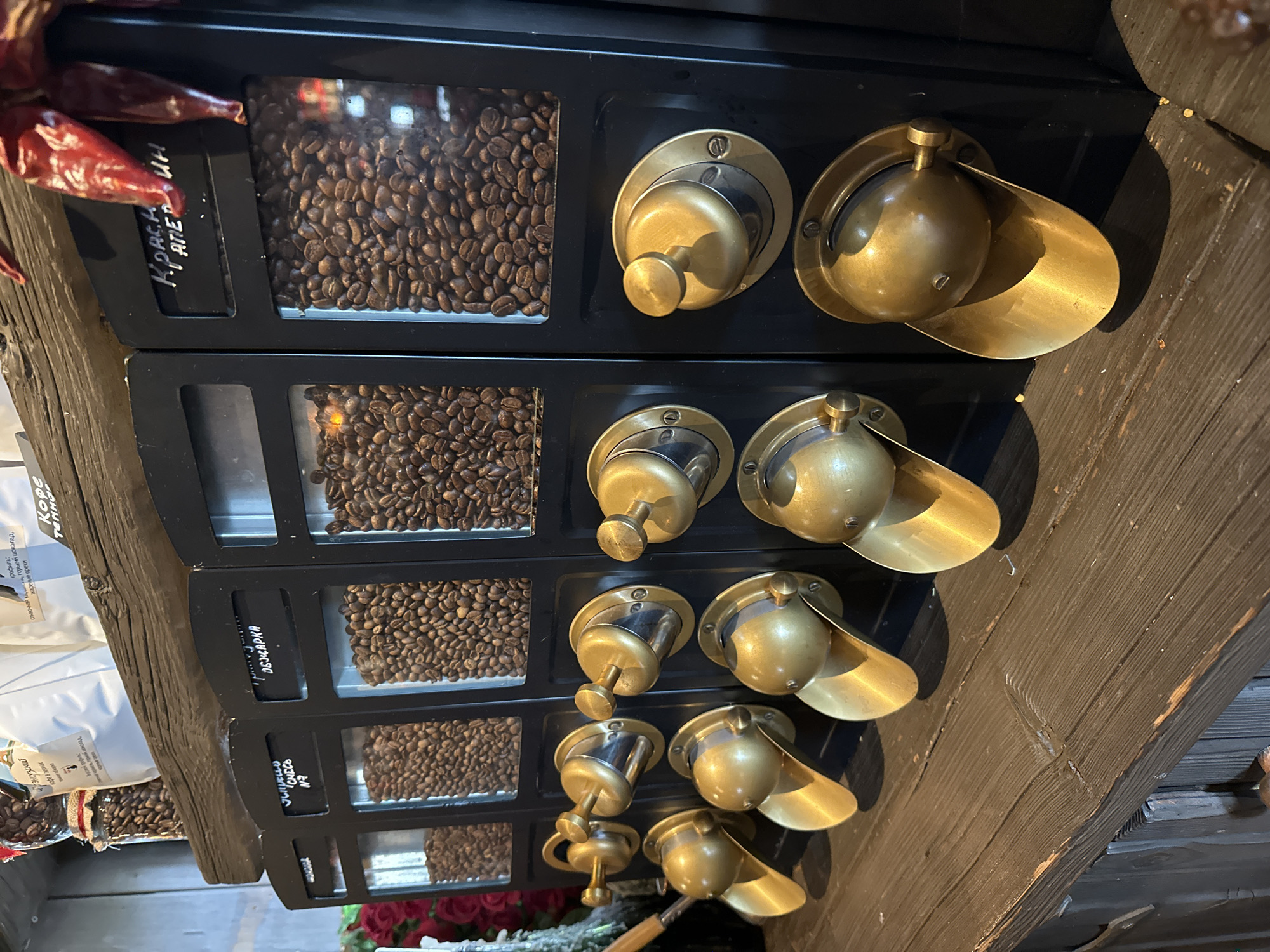 A coffee dispenser in Novoglagolevo, Russia, is depicted in an interior space. The dispenser features four clear containers filled with different roasts of coffee beans, each labeled in Russian. Beside each container is a golden brass dispensing mechanism with a knob and curved spout. To the left of the dispenser are other products, including coffee bags. In the top-left corner, there are dried red chili peppers hanging near the coffee dispenser. The apparatus is mounted on a dark wooden beam.