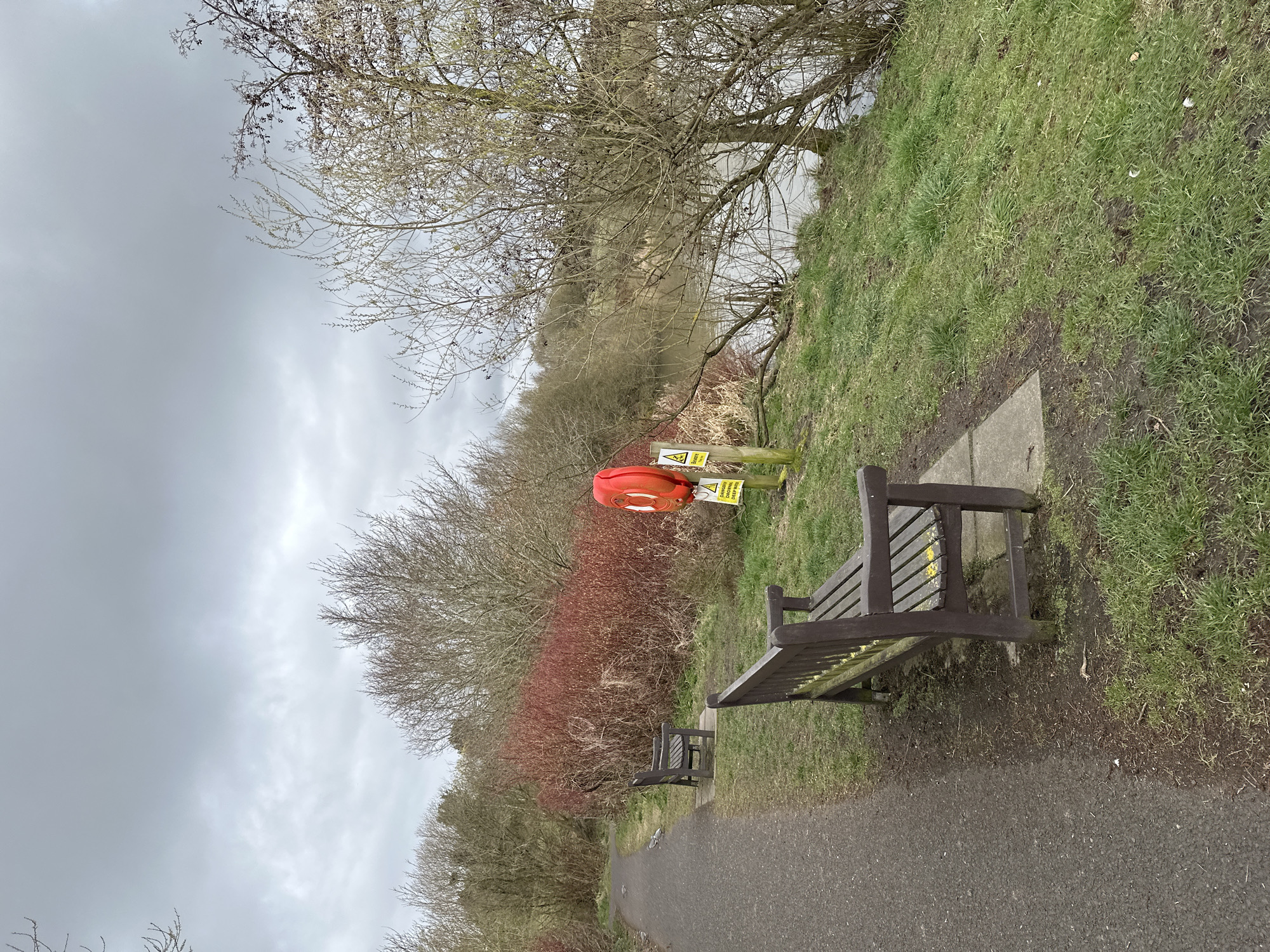 The image shows a park scene in Dunfermline, United Kingdom. A paved path runs along the bottom of the frame, with two dark-stained wooden benches facing towards the right side of the image. A grassy bank rises to the right of the path, meeting the edge of a body of water. A bare tree hangs over the bank. An orange life preserver is mounted on a post behind the first bench, with a yellow "Danger Deep Water" sign attached to the post. The opposite side of the path features a dense collection of bushes, some with reddish-brown foliage. The sky is overcast and gray.