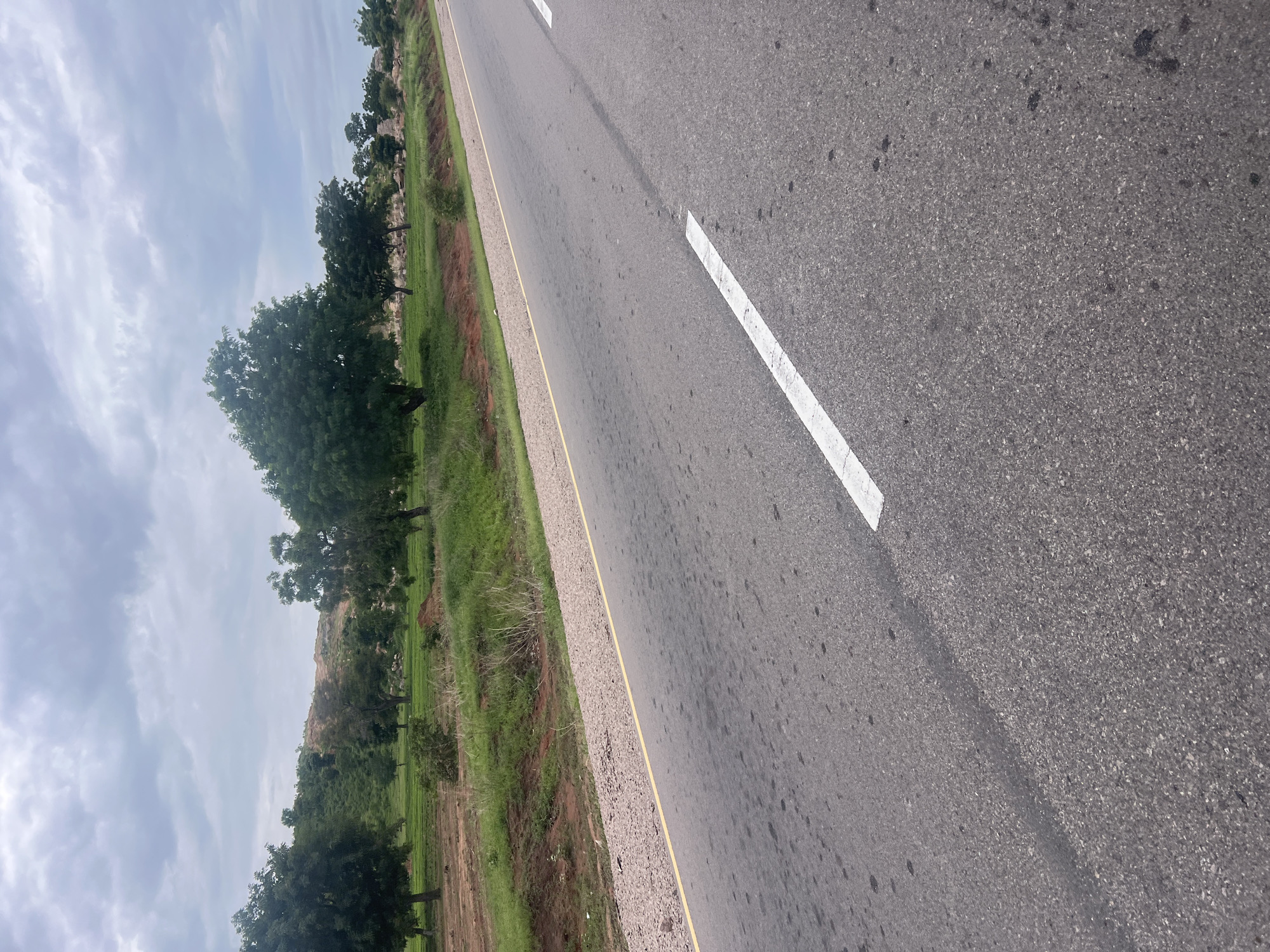 The image displays a roadside scene in Minda, Nigeria. The primary subject is a paved road with a white dashed line marking the lane, indicating it is likely a highway or main road. To the left of the road, there is a strip of green grass and some brown dirt. Further to the left, a line of trees with lush green foliage borders the grassy area. In the background, there are indications of rocky terrain and more trees, suggesting a somewhat rural or semi-urban environment. The sky is cloudy, with a mix of white and gray clouds, hinting at overcast or partly cloudy weather. The lighting suggests it could be daytime, possibly late morning or afternoon. There is no visible text in the image. The perspective is from a low angle, looking across the road and the landscape beside it. The image does not show any people or specific activities, presenting a still, natural landscape with man-made infrastructure.