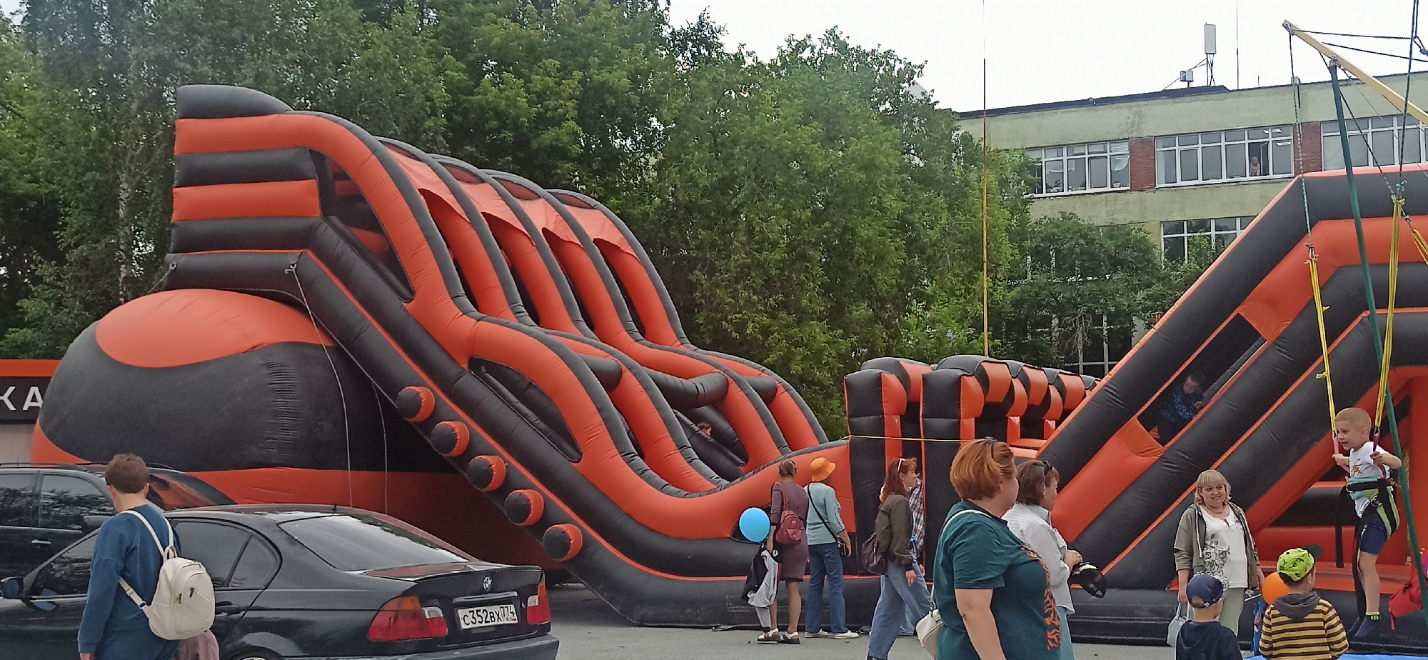 The image shows an outdoor scene featuring a large inflatable obstacle course with orange and black sections. Several people, including children and adults, are present, interacting with the inflatable structure. On the right side, a young boy is being suspended by a bungee harness attached to the inflatable. Other individuals are standing around, observing or participating.
In the background, lush green trees form a dense backdrop, suggesting a natural or park-like setting. Behind the trees, a light-colored building with multiple windows is visible, possibly a school or public facility. The sky is overcast, indicating a cloudy day. The overall atmosphere appears to be one of recreational activity, with families enjoying themselves.
To the left of the inflatable, several parked cars are visible, including a black BMW with a license plate that reads "C352BX 774". A woman wearing a blue sweater and a white backpack is walking away from the camera, passing by the cars. Another woman with an orange hat is standing near the inflatable.
The scene is likely taking place during the daytime, given the ambient light. The presence of the inflatable structure and the activities of the people suggest an event or a public playground setup. The setting seems to be a paved area, possibly a parking lot or an open space near a recreational facility. There are no strong indicators of specific emotions beyond general engagement and enjoyment of the outdoor activities.