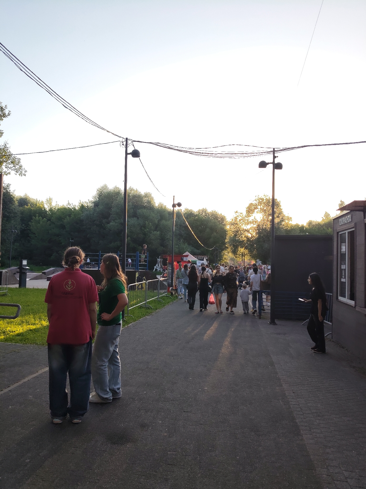 The image depicts an outdoor scene in the city of Chekhov, Russia, likely at a park or event space. In the foreground, two individuals, one wearing a red polo shirt and the other a green t-shirt, are facing away from the camera and conversing. They are standing on an asphalt pathway. Beyond them, a crowd of people is walking along the same pathway, some with shopping bags, suggesting a market or festival. In the background, there are trees and a structure that appears to be a booth or kiosk with the word "ОХРАНА" (security) visible. The lighting suggests it is late afternoon or early evening, with the sun casting long shadows. Power lines are visible crisscrossing the sky.